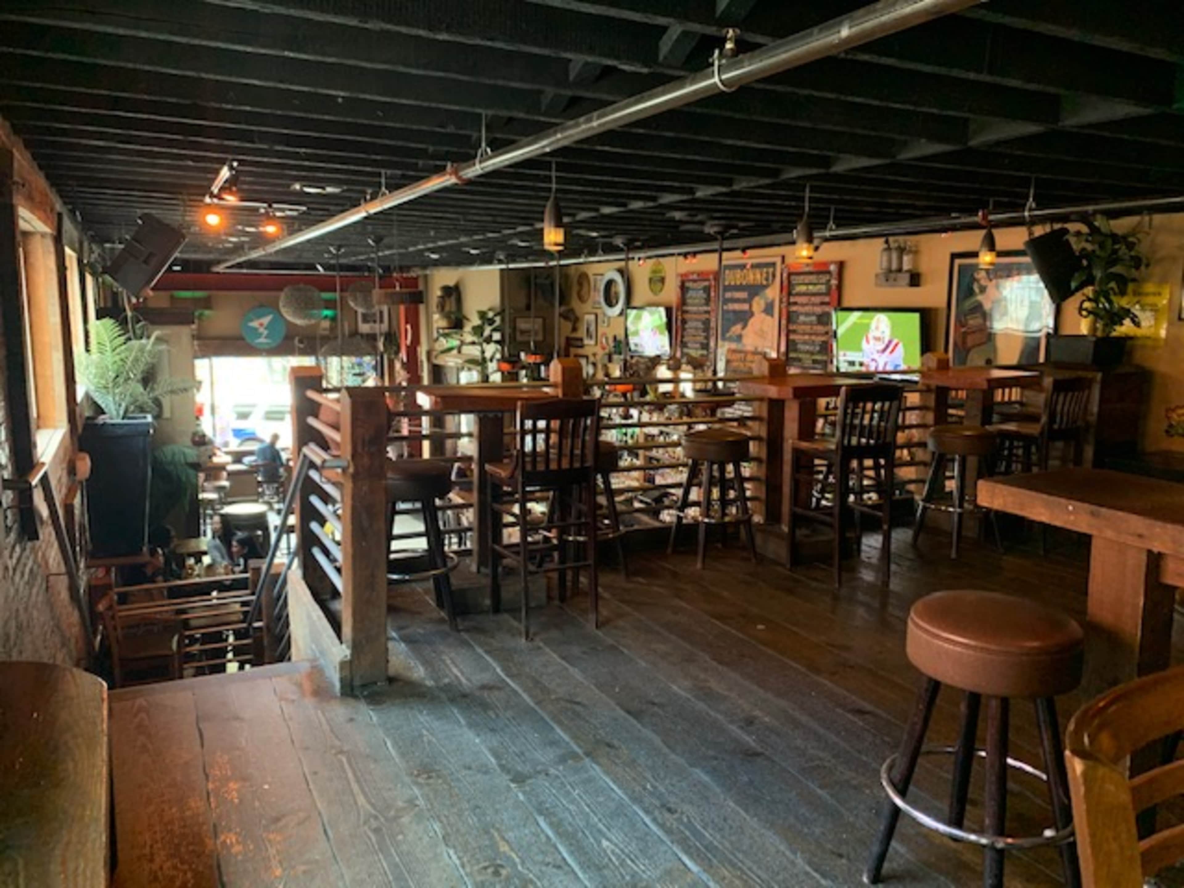 A rustic bar area with wooden furniture, including tables and stools, illuminated by warm lighting and featuring a variety of decorations on the walls.