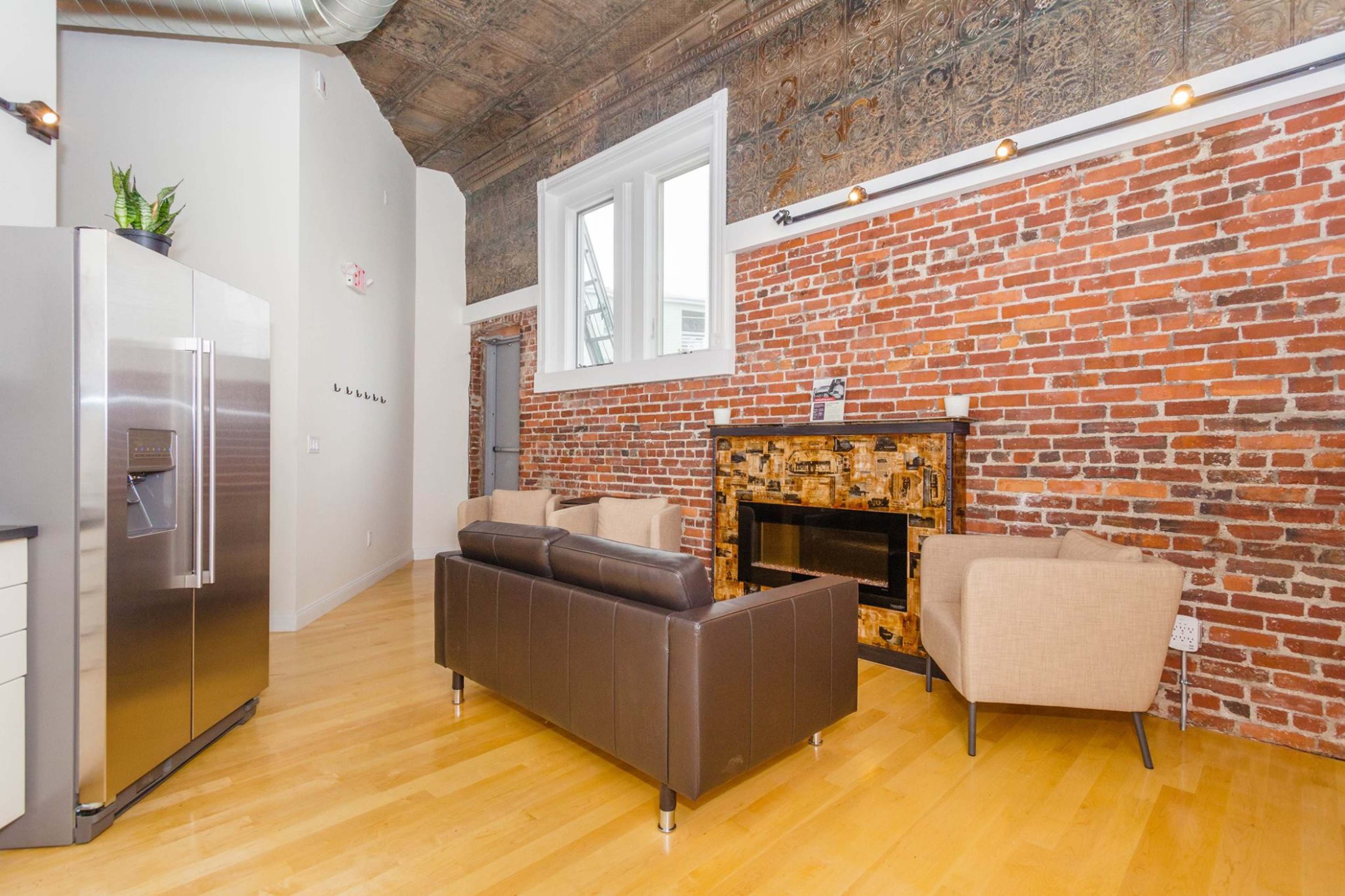 A modern living space with exposed brick walls, a sleek refrigerator, and a cozy seating arrangement near a decorative fireplace.