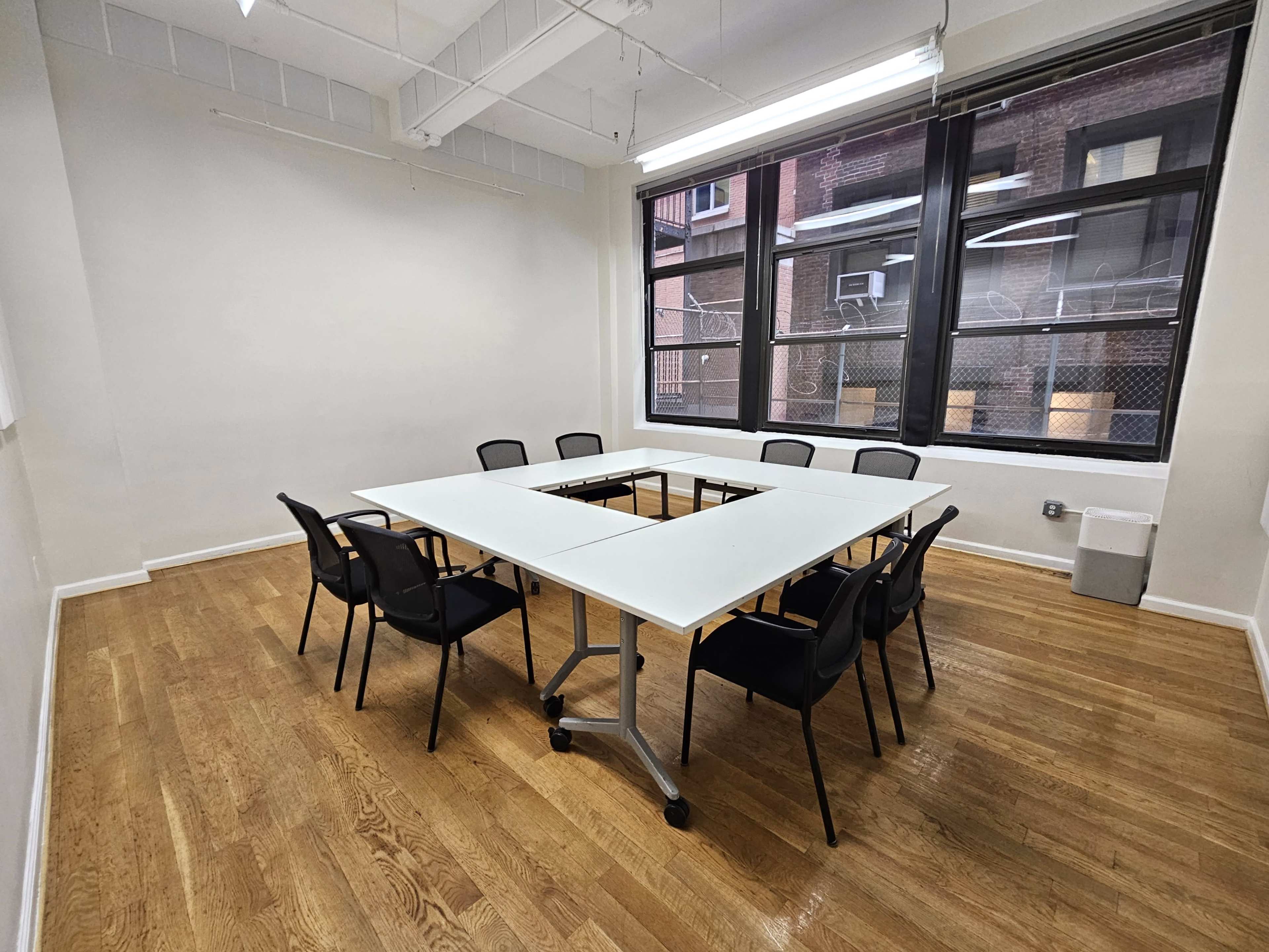 A rectangular conference table with eight black chairs is placed in a well-lit meeting room featuring large windows.