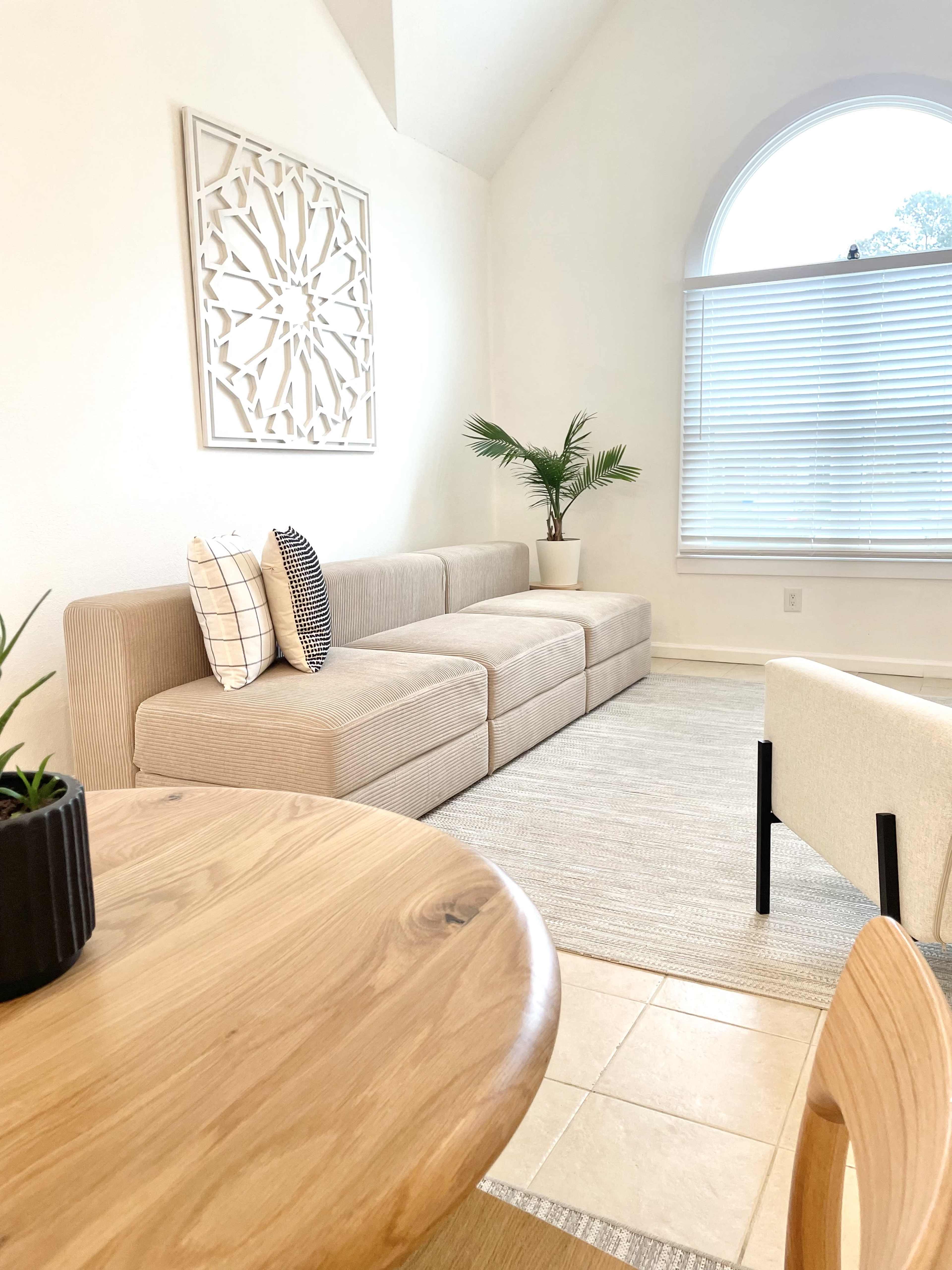 A modern living room with a beige sectional sofa, a round wooden dining table, a potted plant, and a large window covered by sheer blinds.