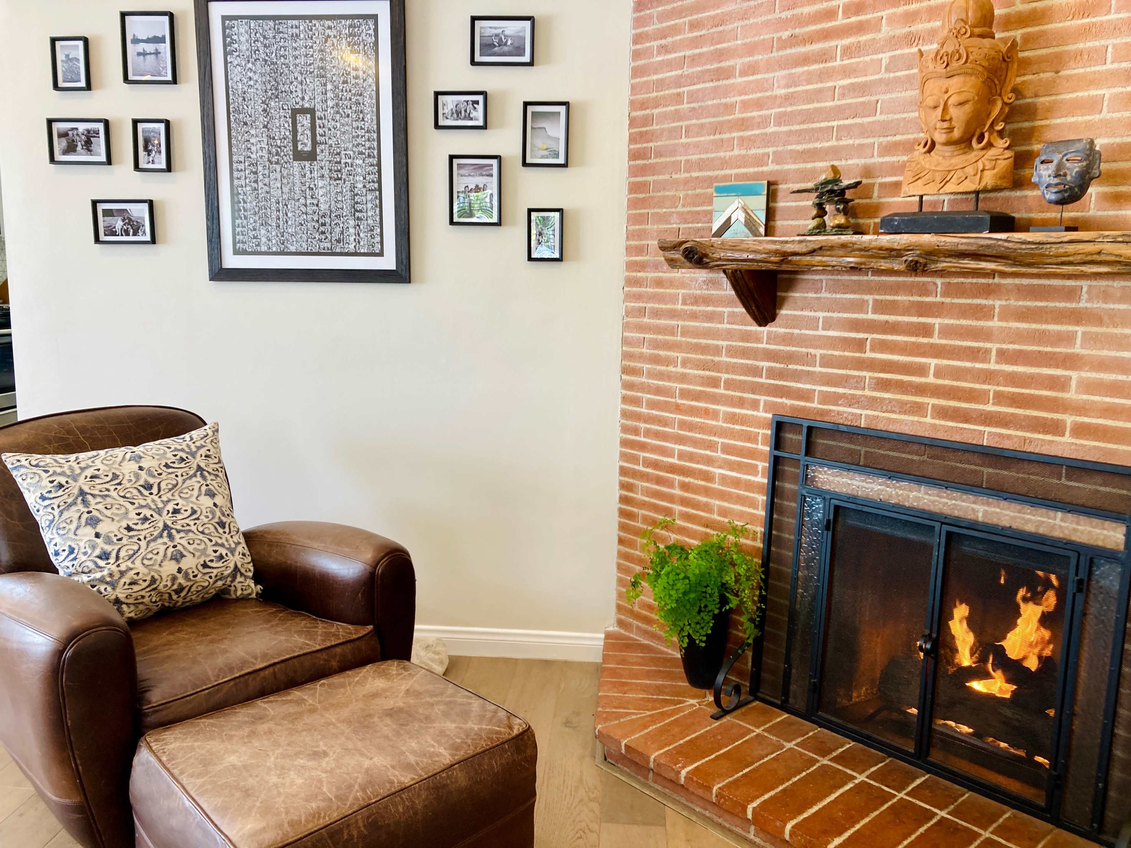 A brown leather chair with a matching ottoman sits beside a brick fireplace adorned with a shelf displaying decorative items and framed photos on the wall.