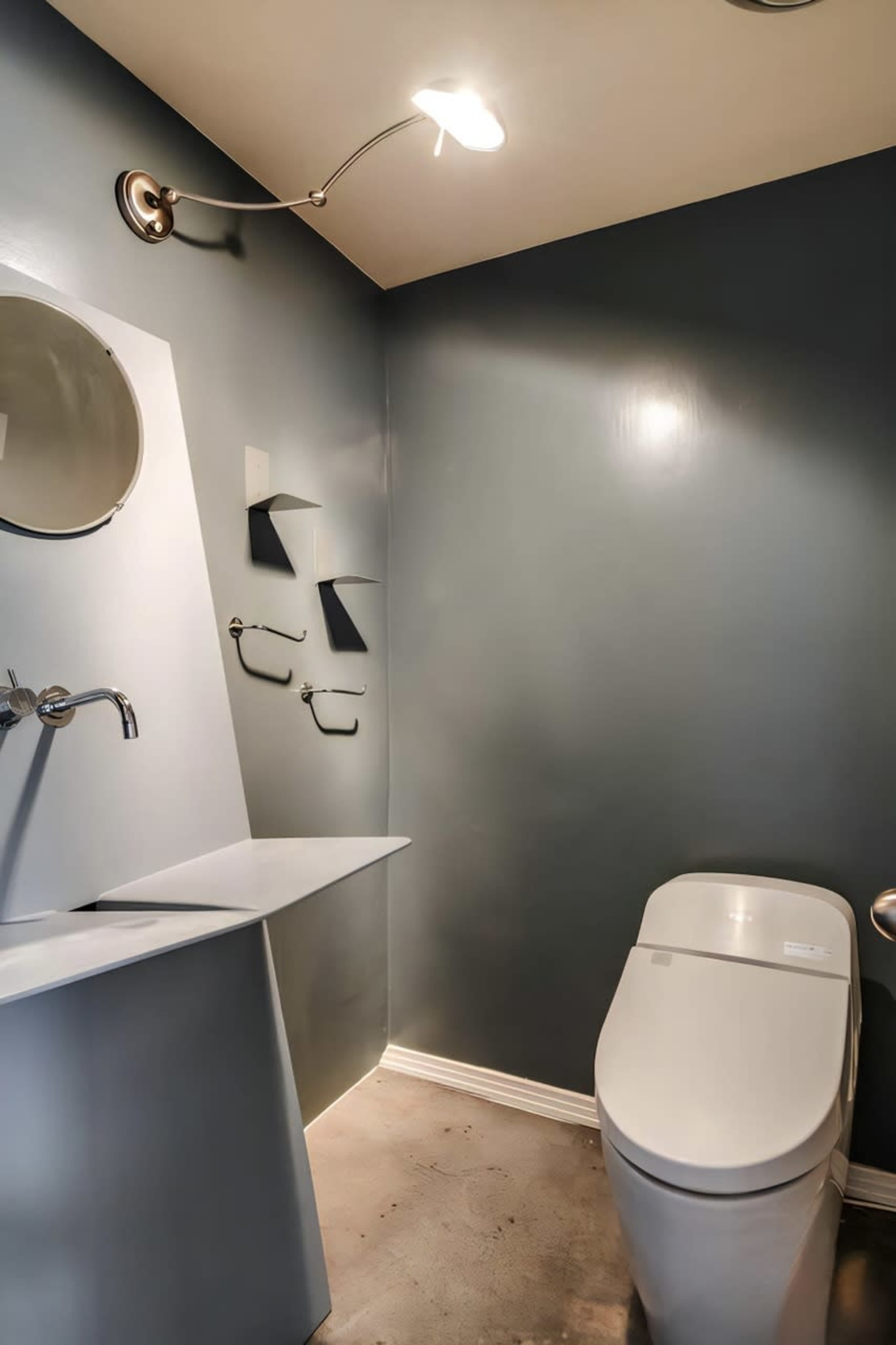 The image shows a modern bathroom with a sleek white toilet, a curved sink counter, and a circular mirror against a gray wall.