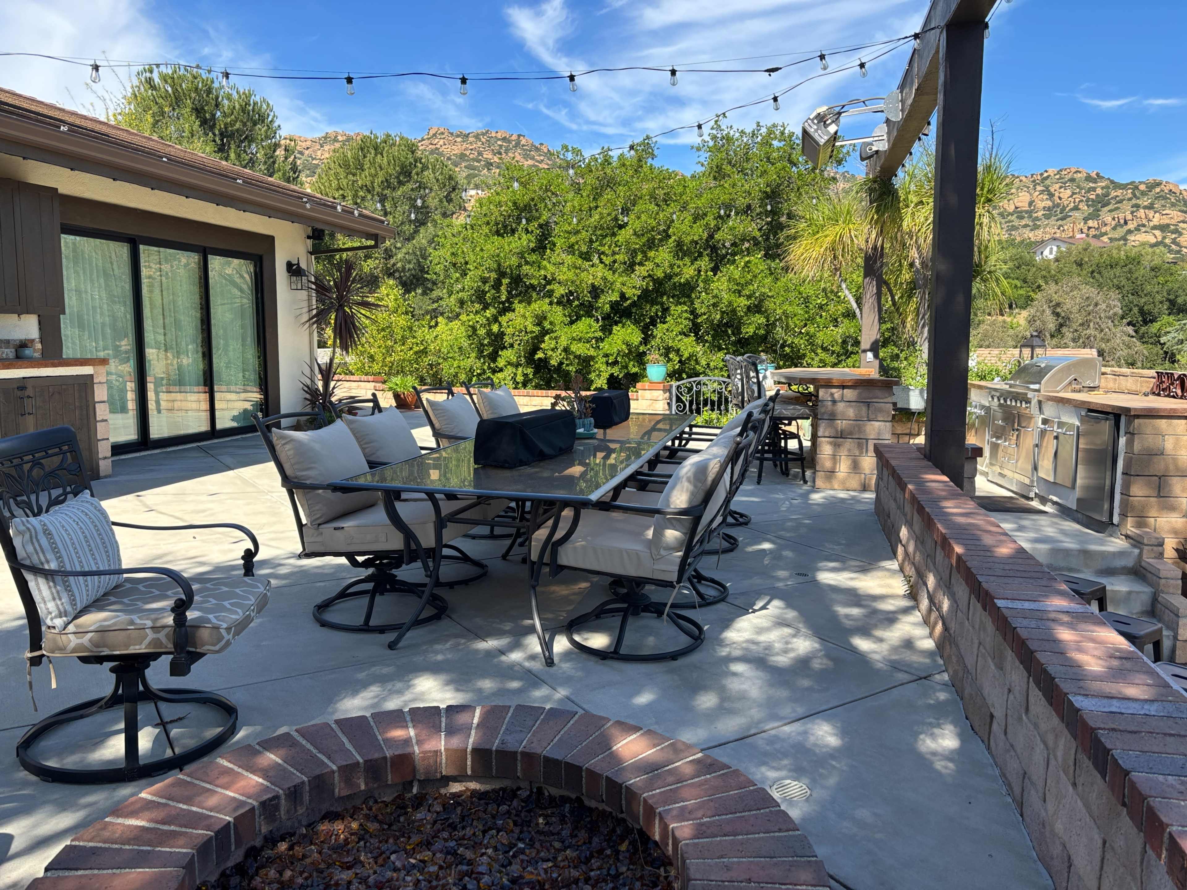 The image shows a patio area with a large dining table surrounded by chairs, a barbecue grill, and trees in the background under a blue sky.