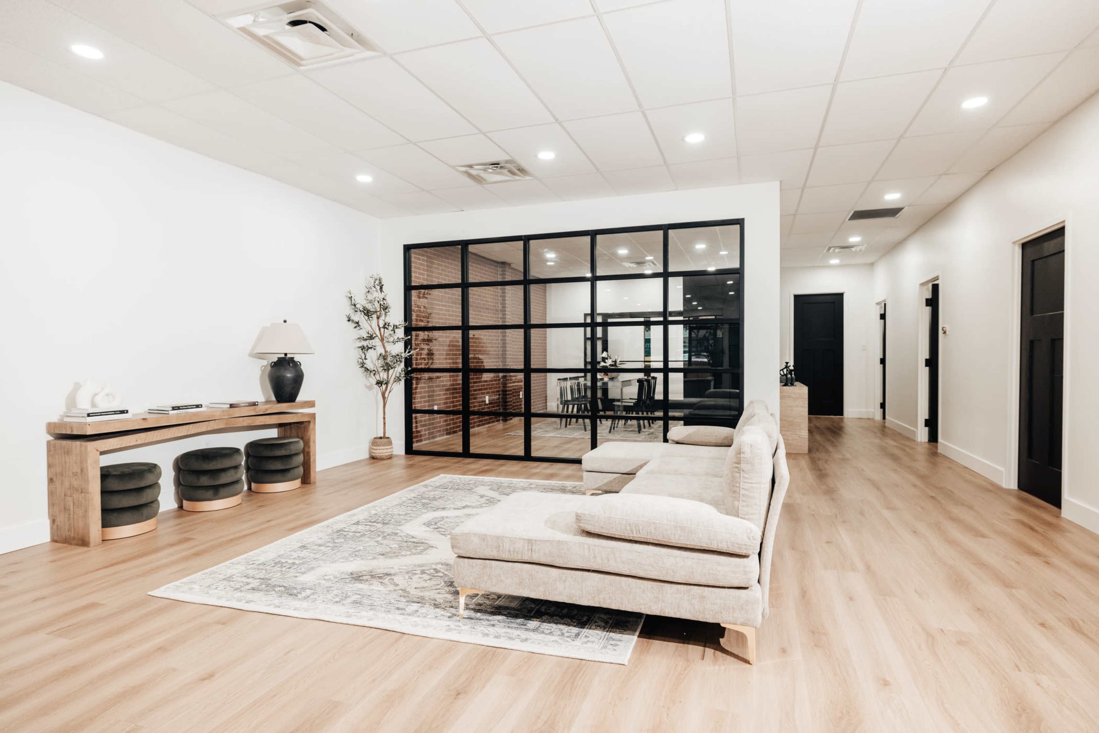 A spacious room features a modern beige sectional sofa, a wooden console table, and a glass-enclosed meeting space in the background.