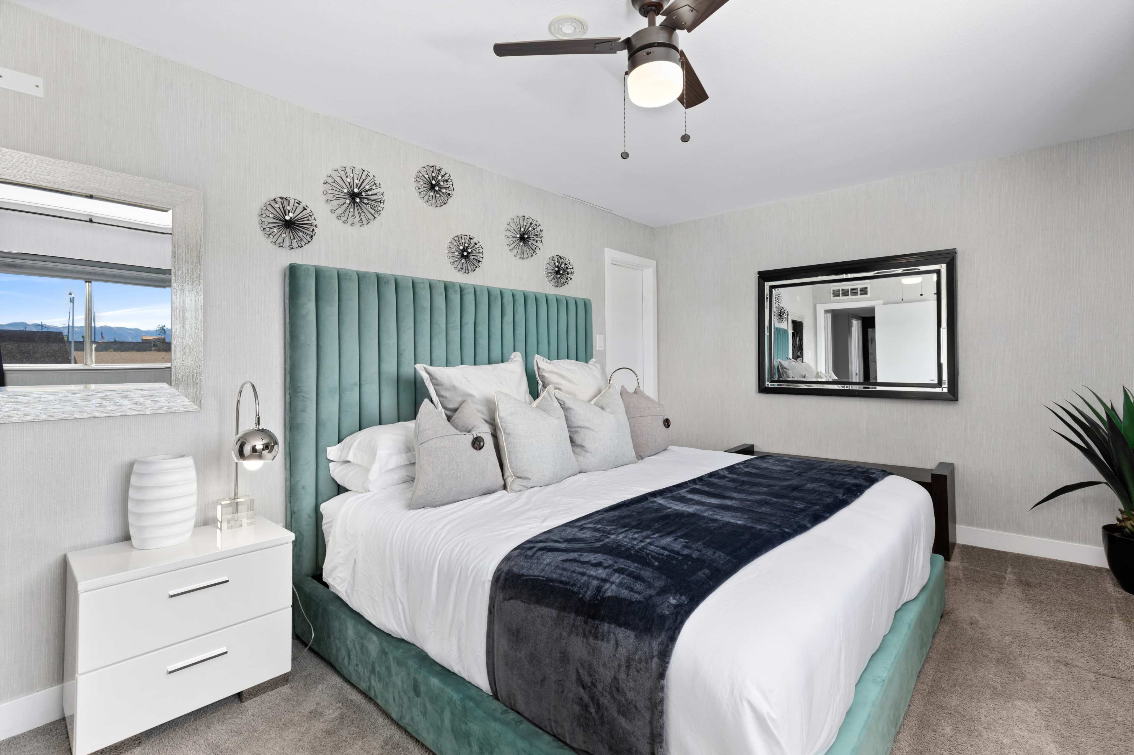 A modern bedroom features a large upholstered bed with a dark gray throw, accent pillows, and geometric wall decor, complemented by a nightstand and a mirror.