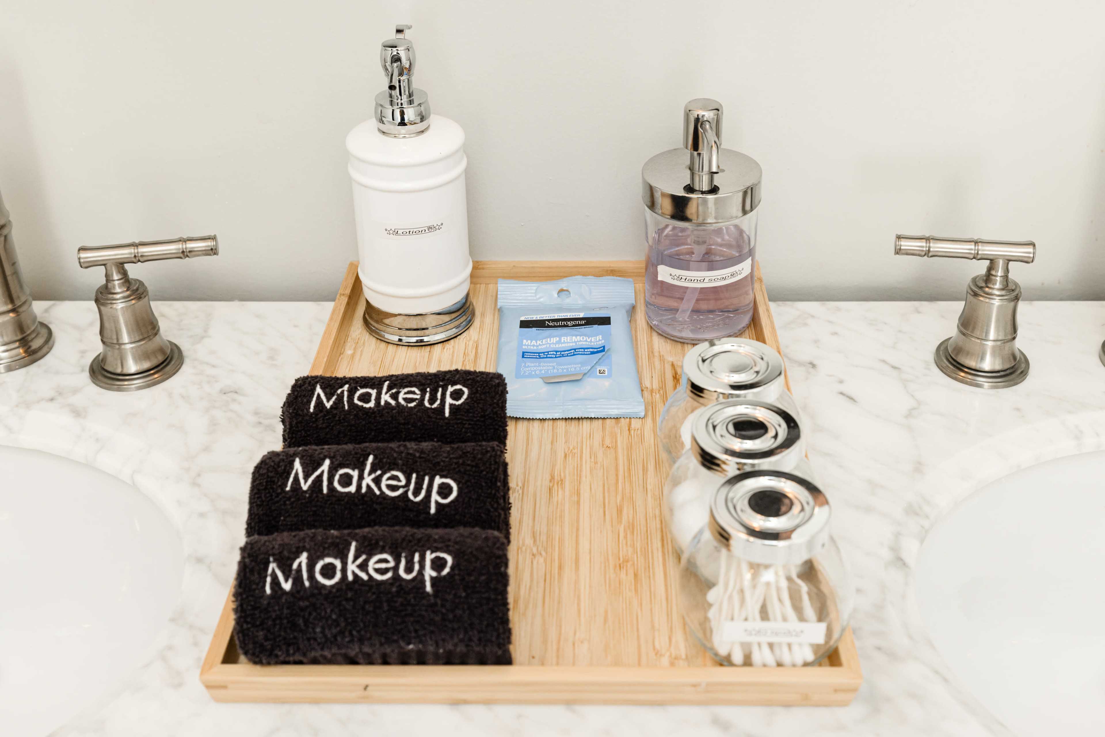A wooden tray on a marble countertop holds labeled towels, skincare products, and containers for cotton swabs.