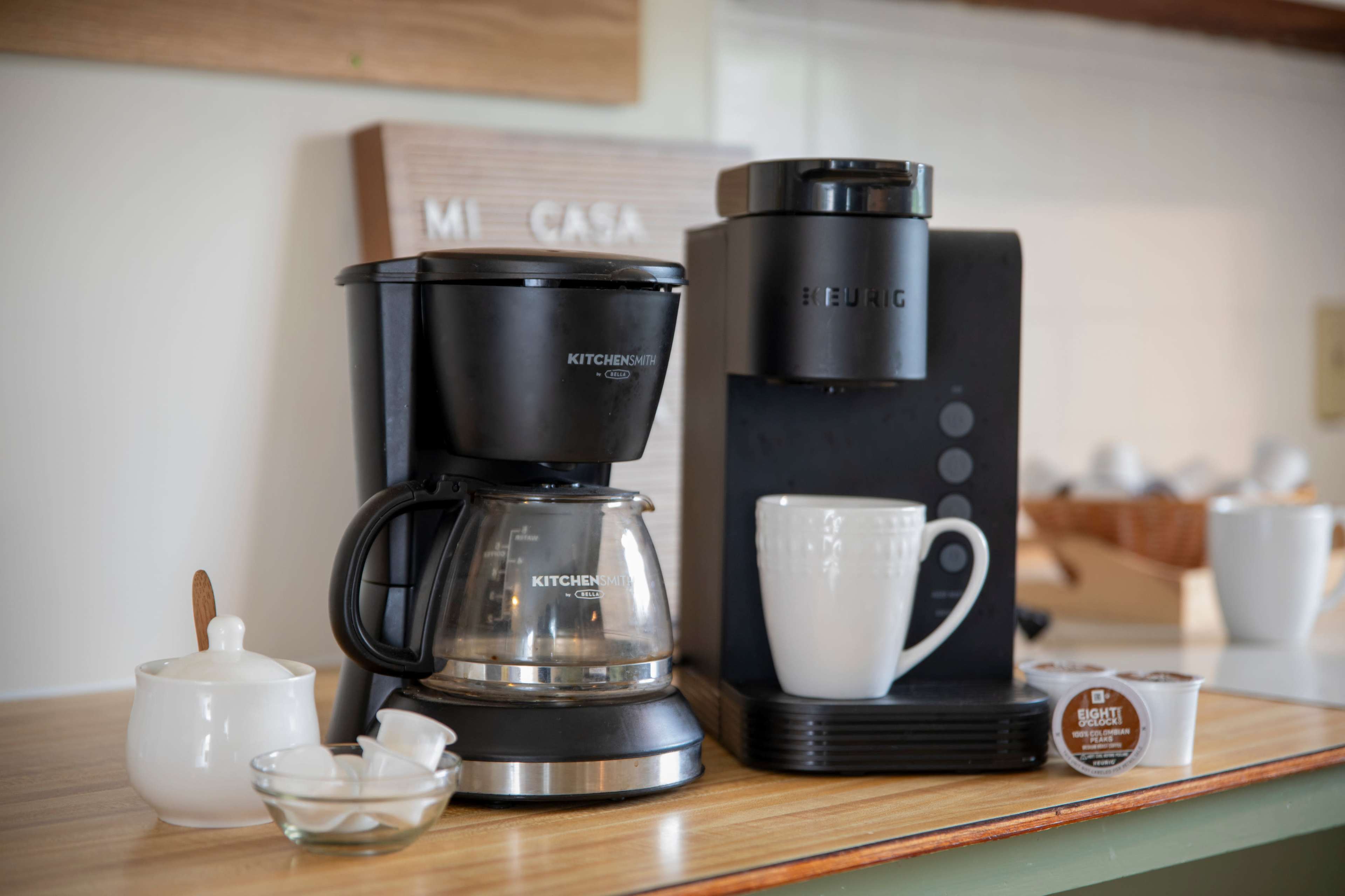 A traditional drip coffee maker and a single-serve coffee machine are displayed on a kitchen counter beside a white cup and a sugar container.
