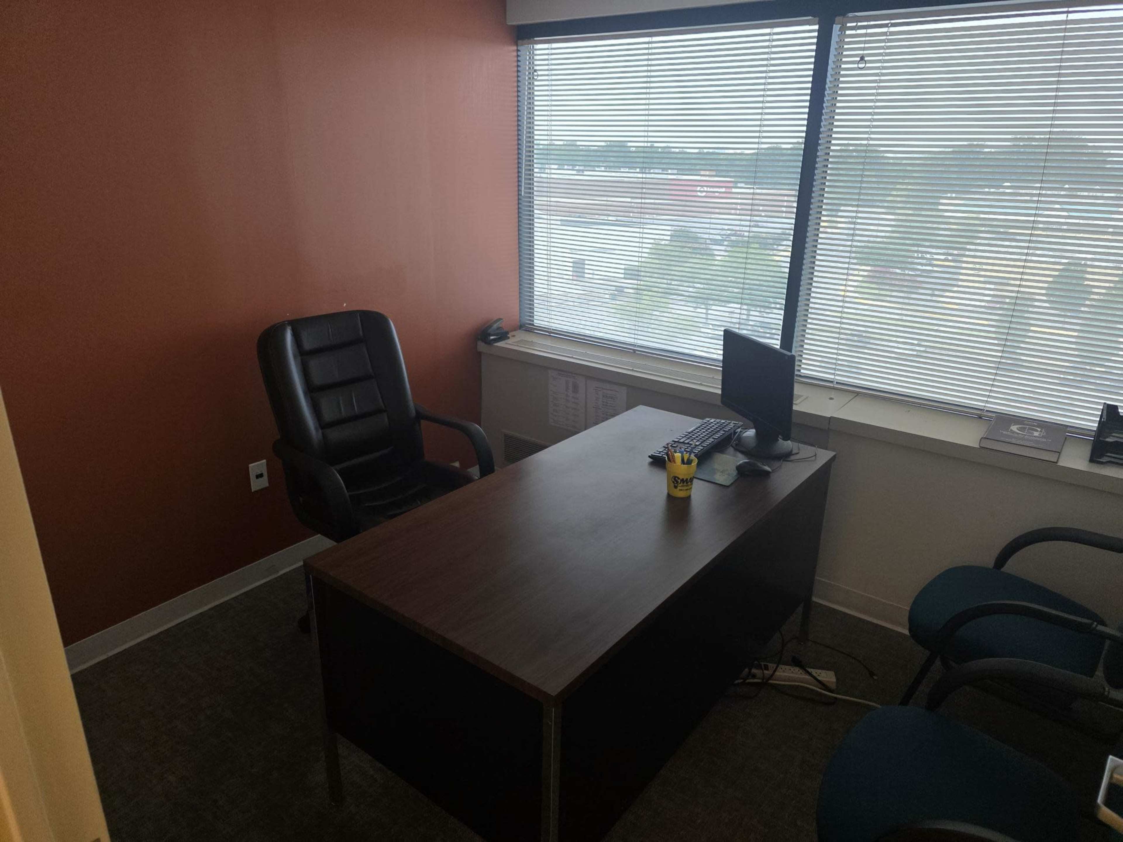The image shows a small office with a wooden desk, a black office chair, a computer on the desk, and large windows with blinds overlooking an exterior view.