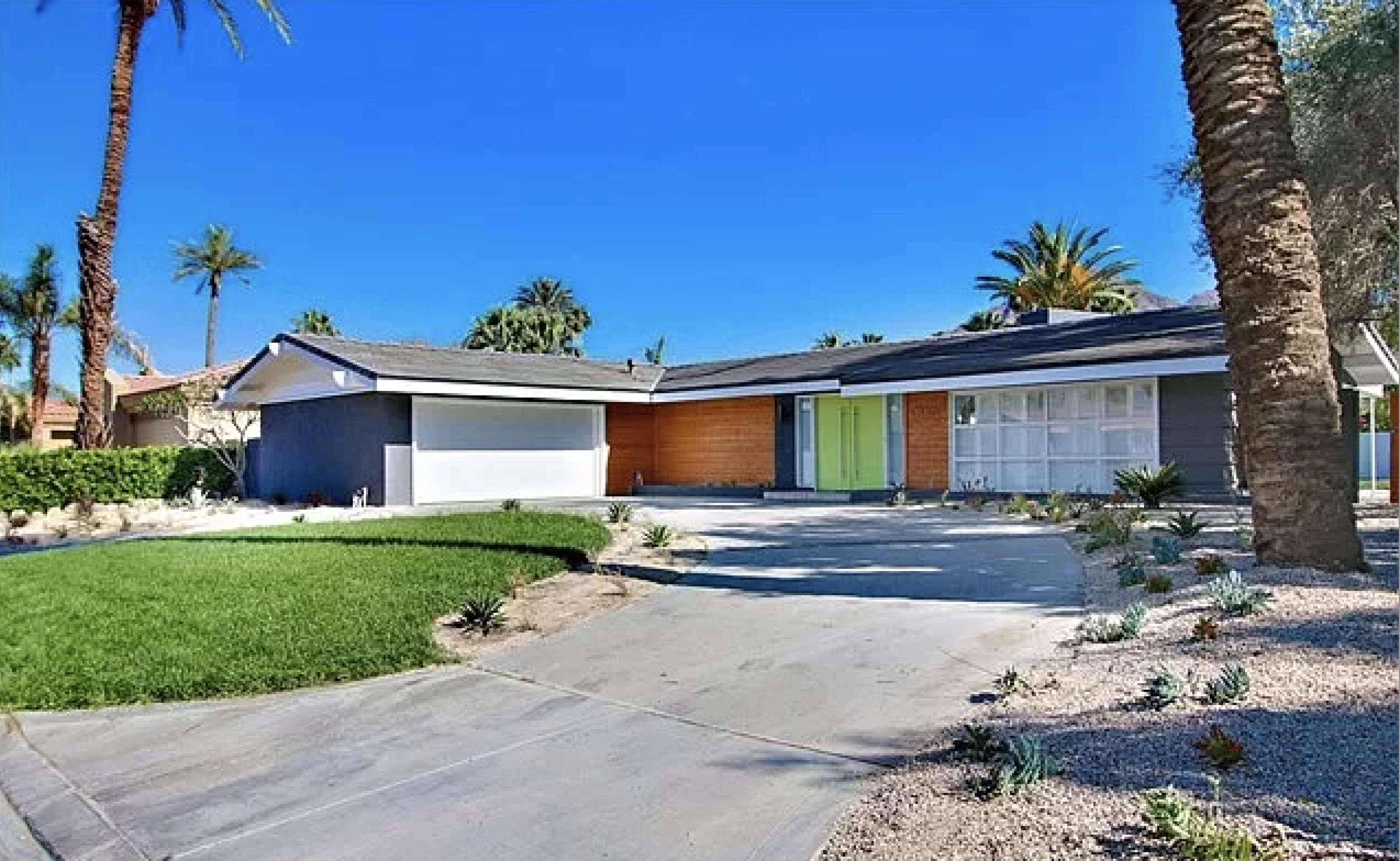 A modern single-story home features a driveway leading to a garage and a bright green front door, surrounded by palm trees and landscaped greenery.