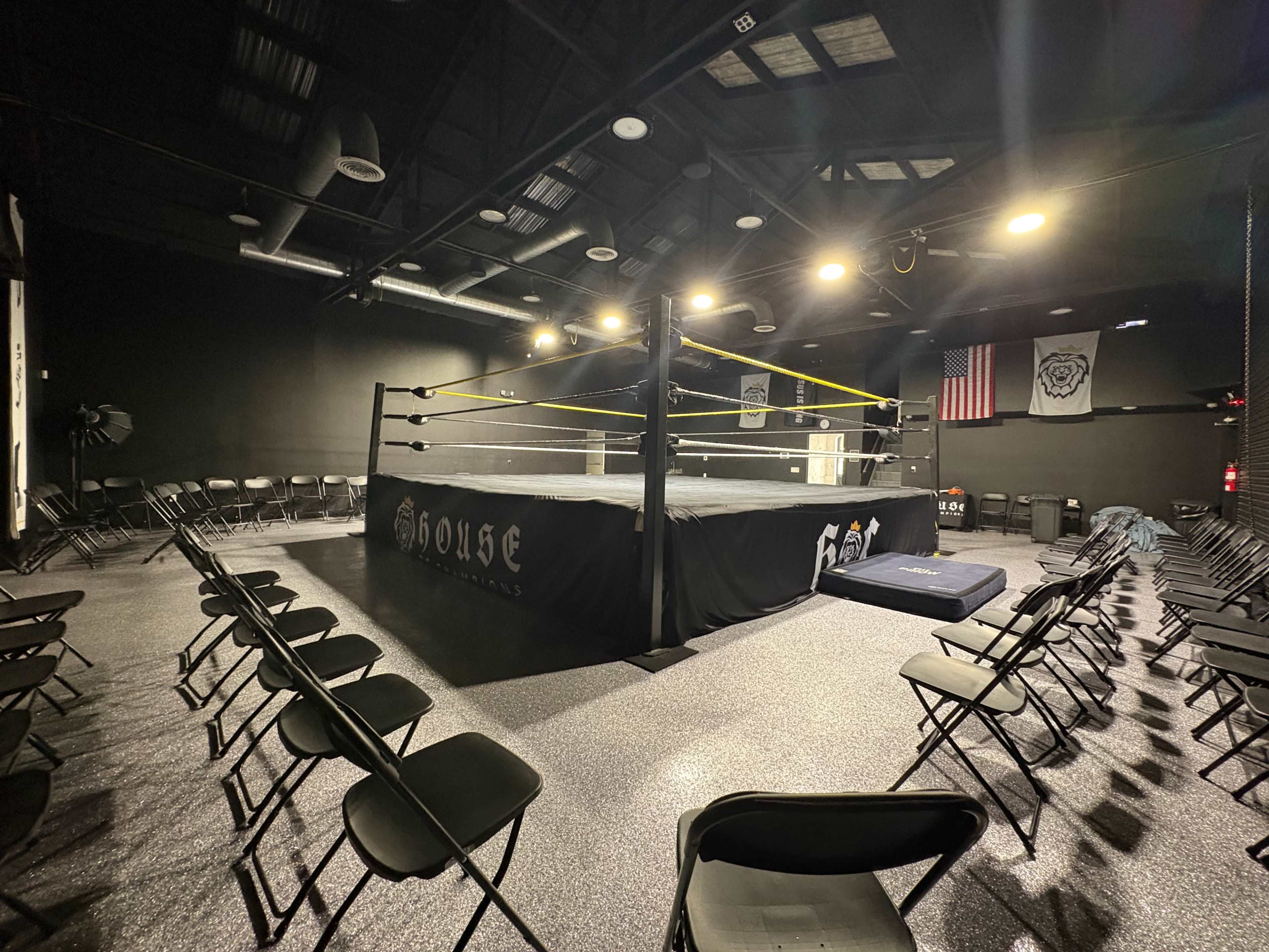The image depicts an empty wrestling ring surrounded by black folding chairs, with dim lighting highlighting the area inside a training facility.