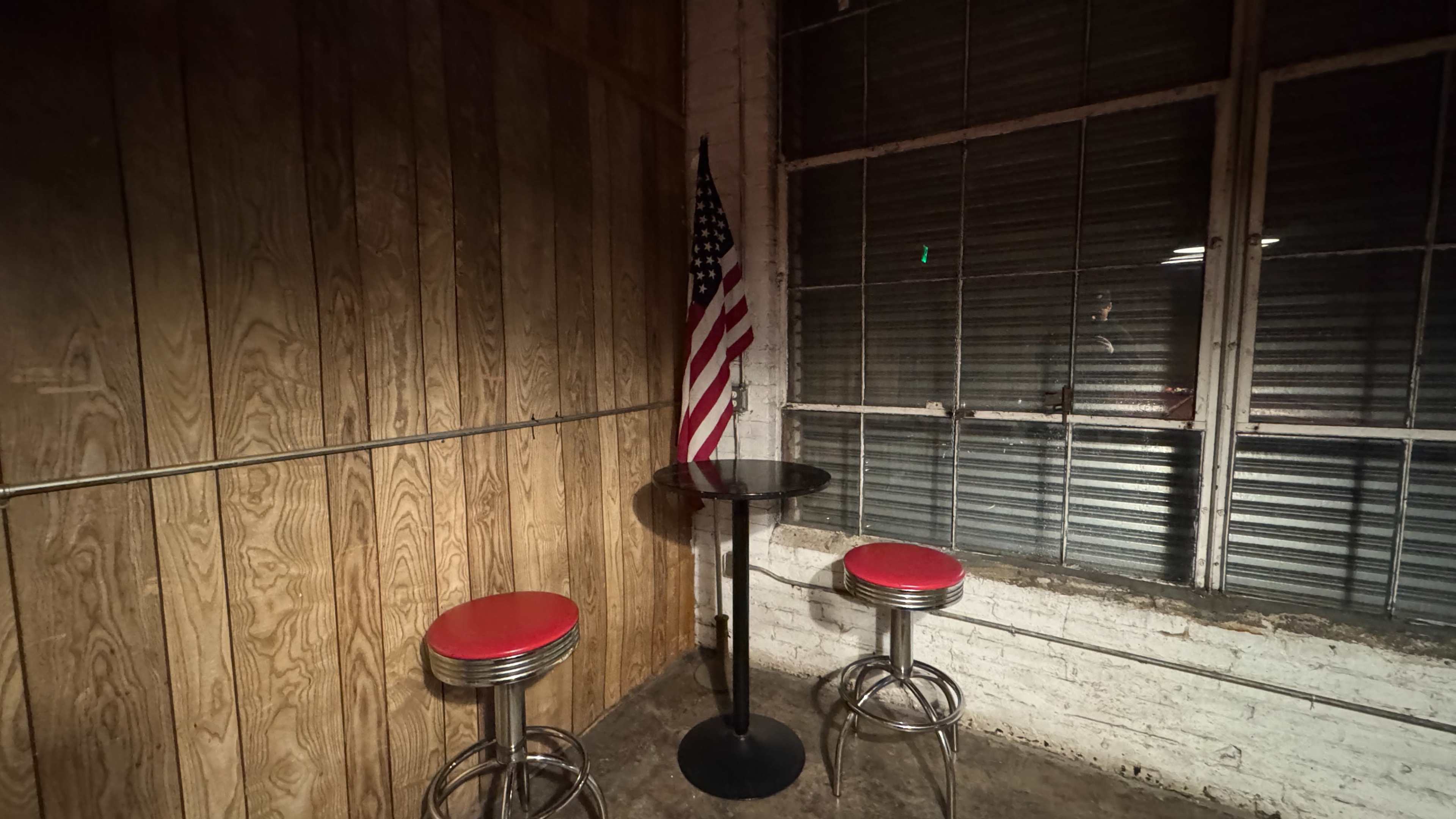 A small table with two red bar stools sits against a wooden wall, beside an American flag and a large window with closed blinds.