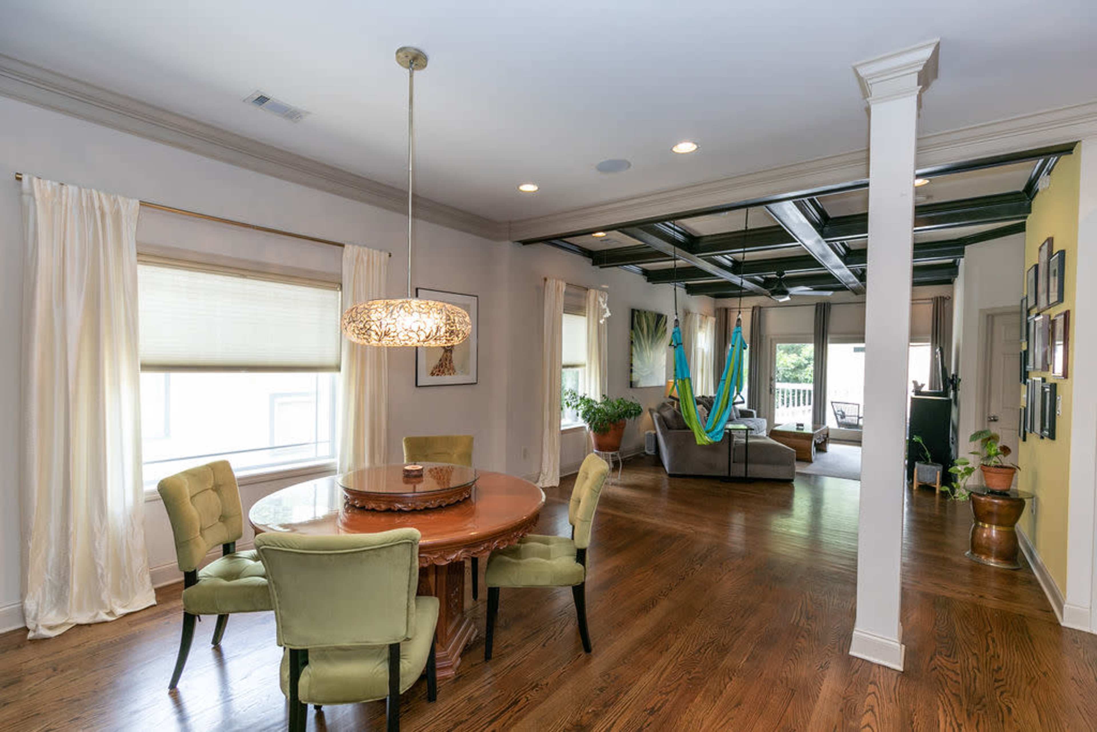 A spacious living area with a round dining table, four green chairs, and a view of a sunlit room featuring multiple hammocks and large windows.