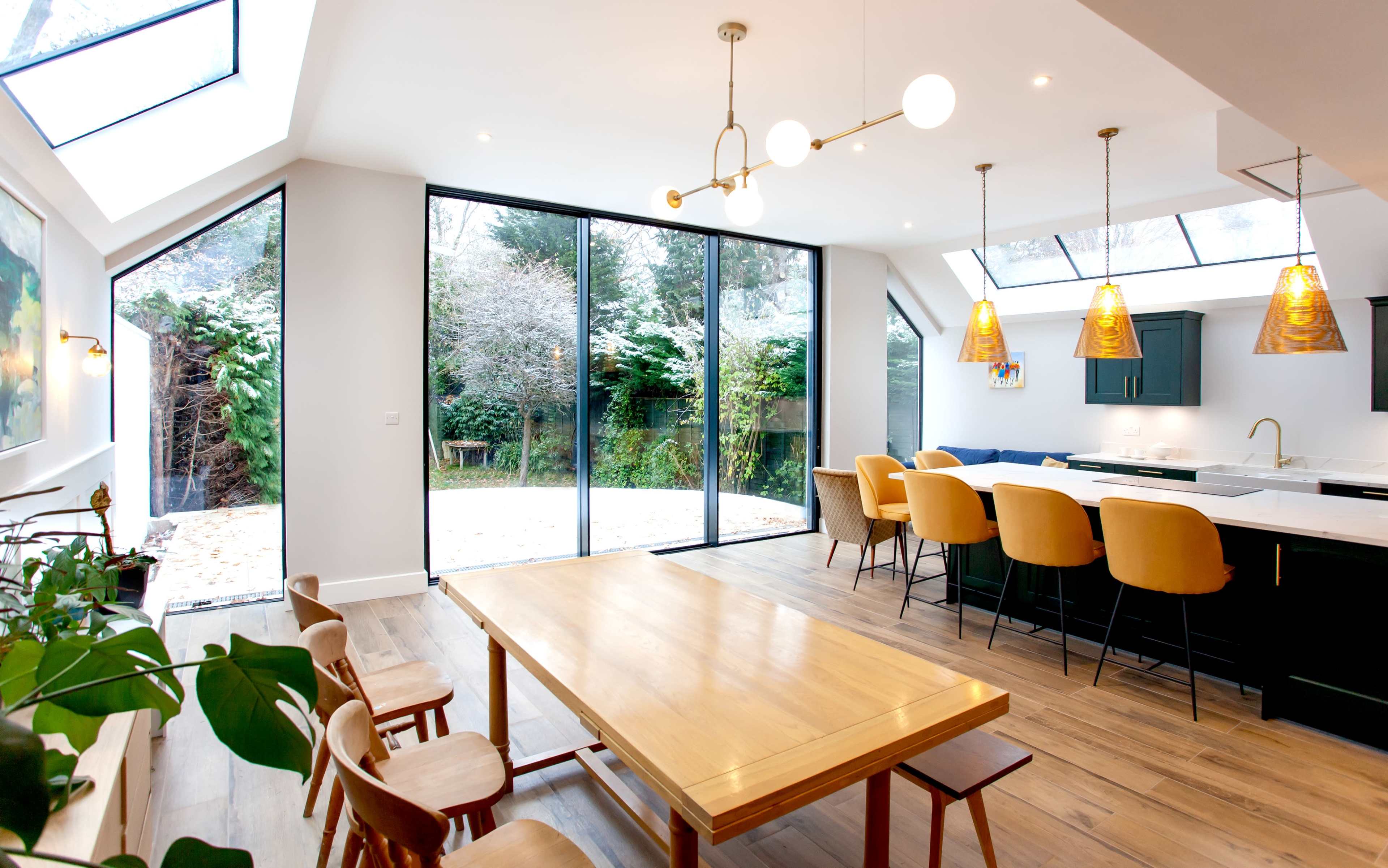 A modern kitchen-dining area features a large wooden table, bar seating, and large glass doors leading to a garden.