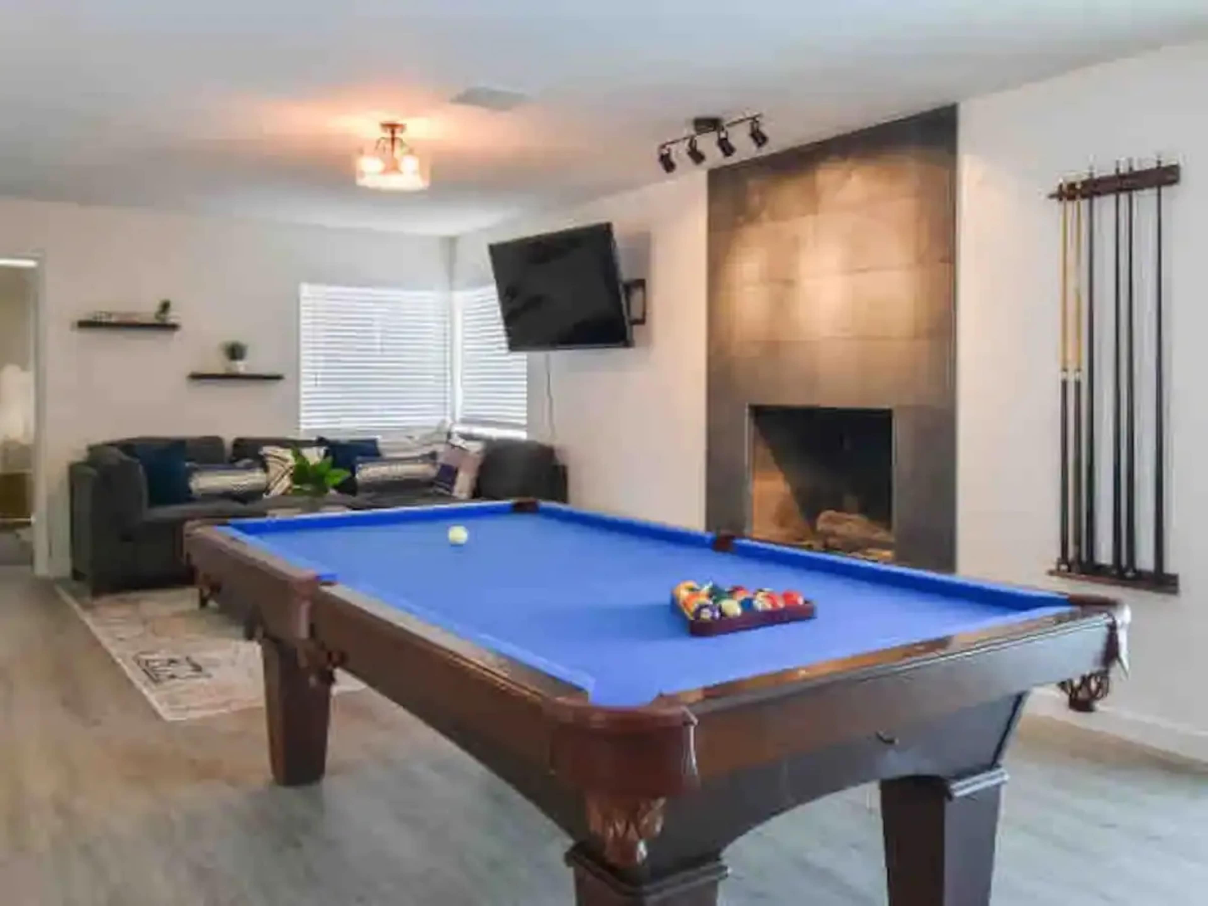 The image shows a game room featuring a blue pool table, a fireplace, a mounted television, and a seating area with a couch.