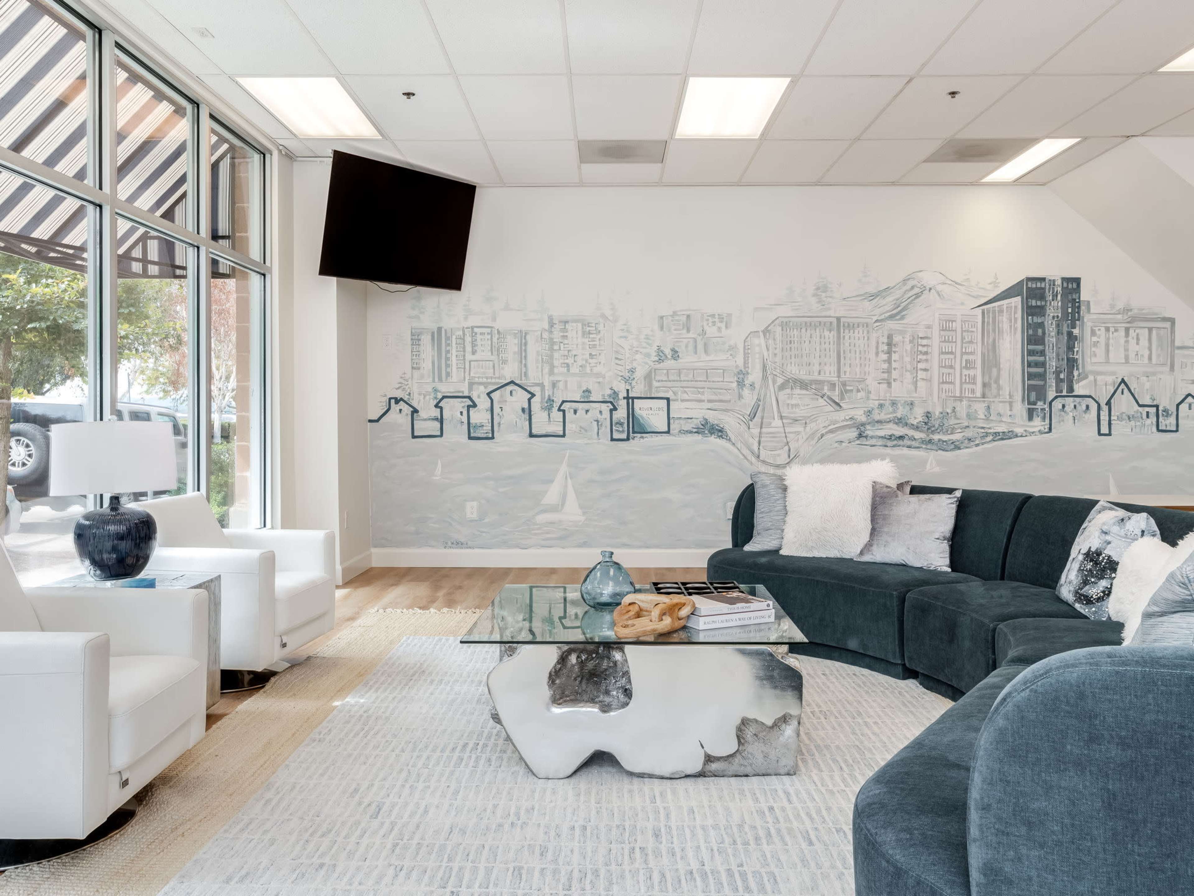 The image depicts a modern lounge area featuring a sectional couch, white armchairs, a glass coffee table, and a mural of a cityscape on the wall.