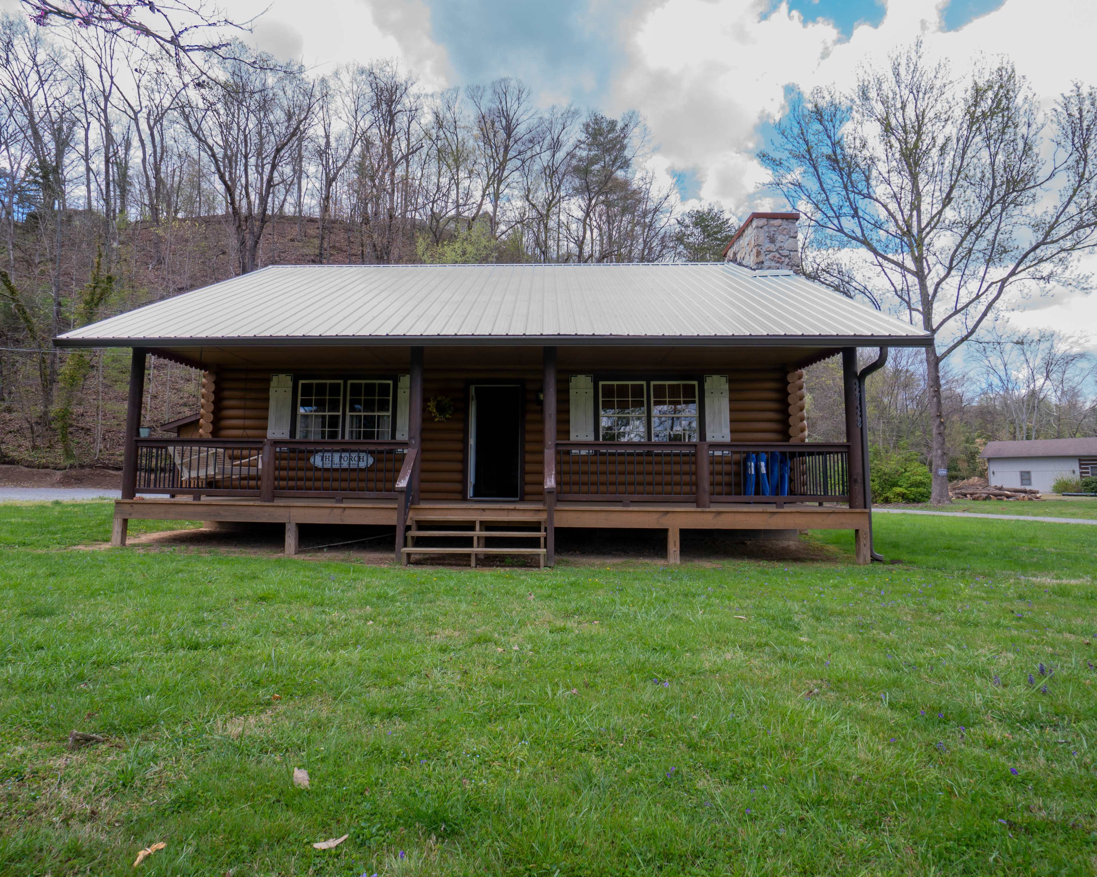 A log cabin with a metal roof features a front porch and is surrounded by green grass and trees.