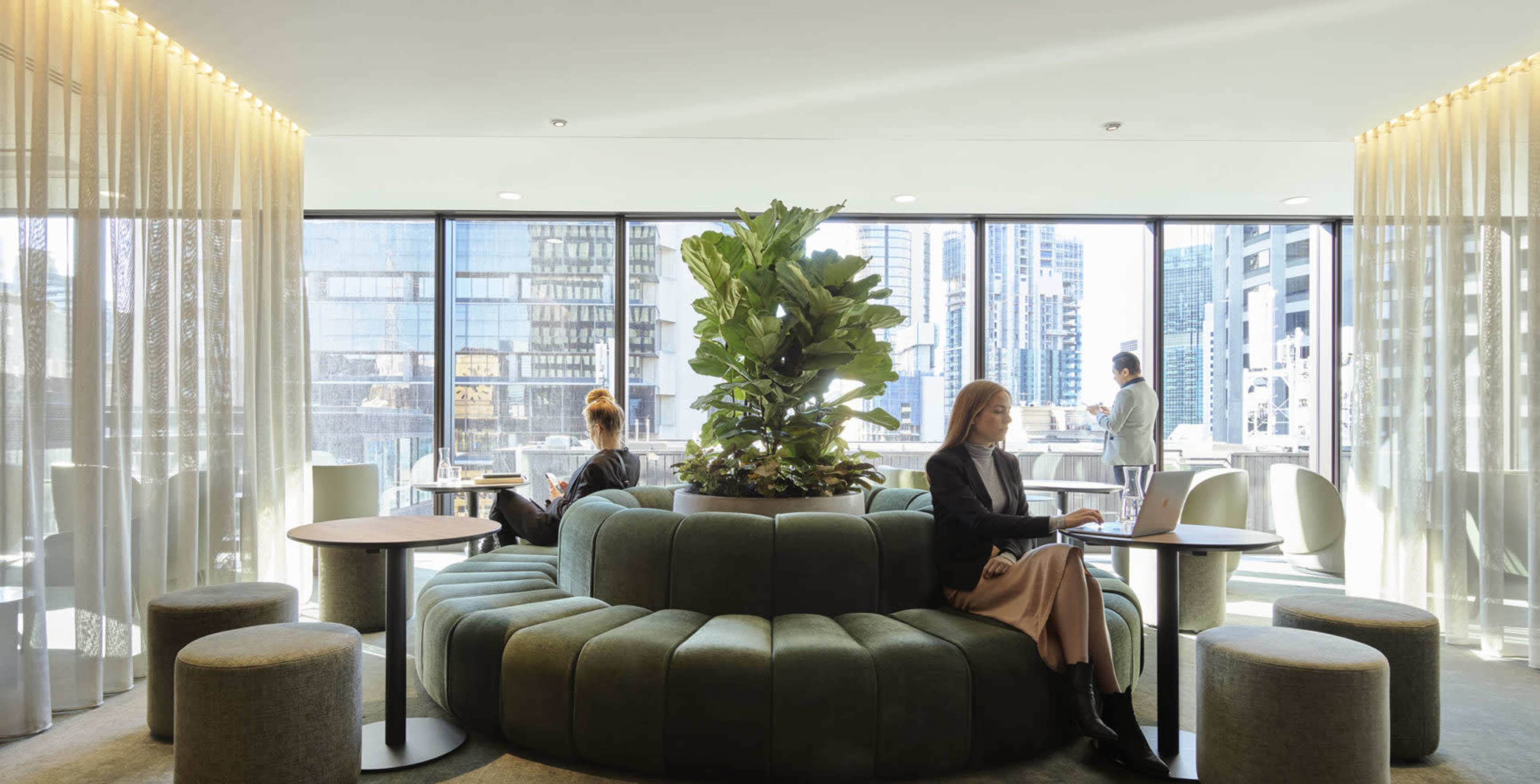 A modern workspace with a circular green seating area, large windows featuring a cityscape, and individuals working on laptops.