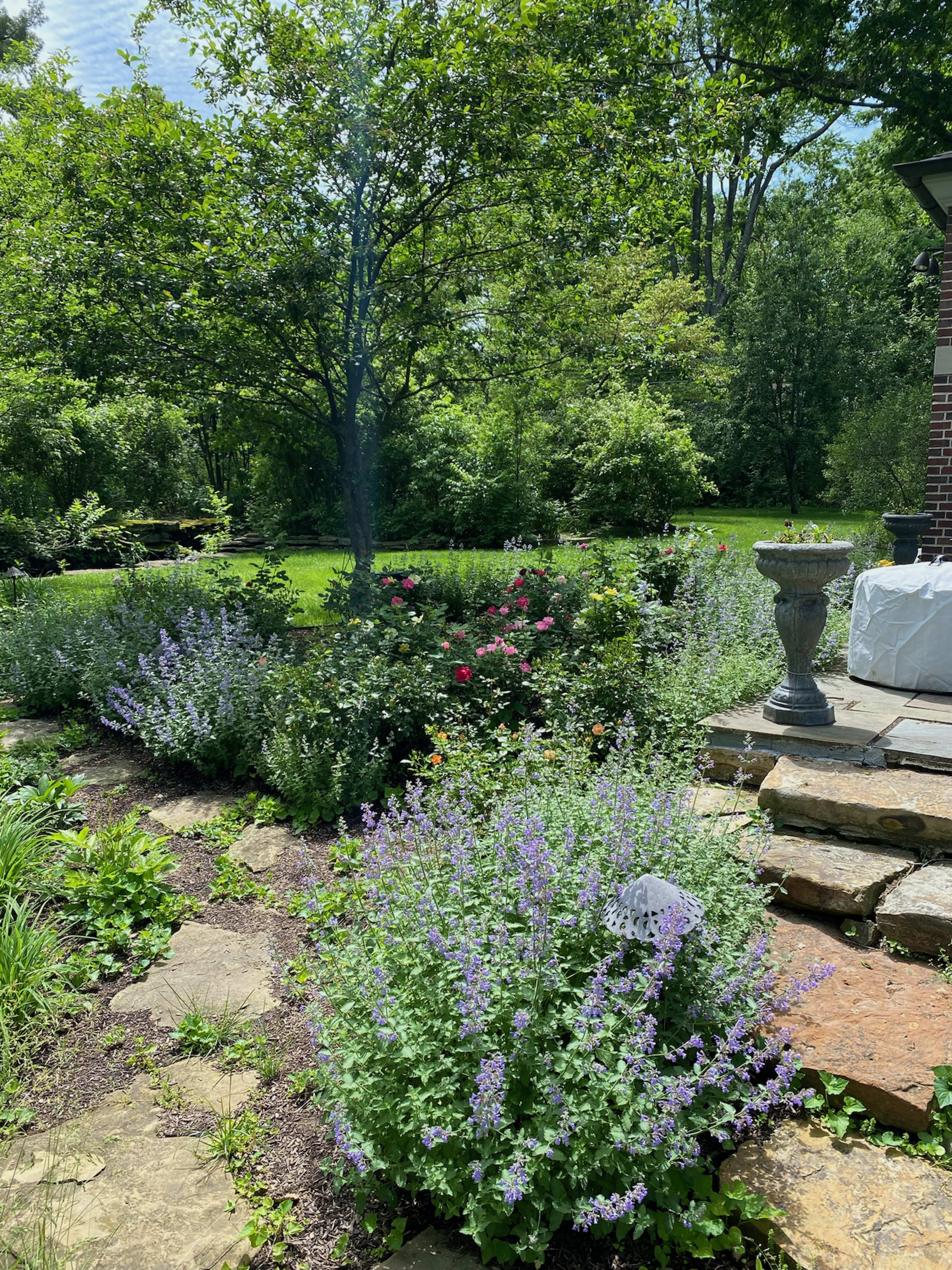 A garden area features a variety of flowering plants surrounded by stone pathways and a decorative urn, with trees and grass in the background.