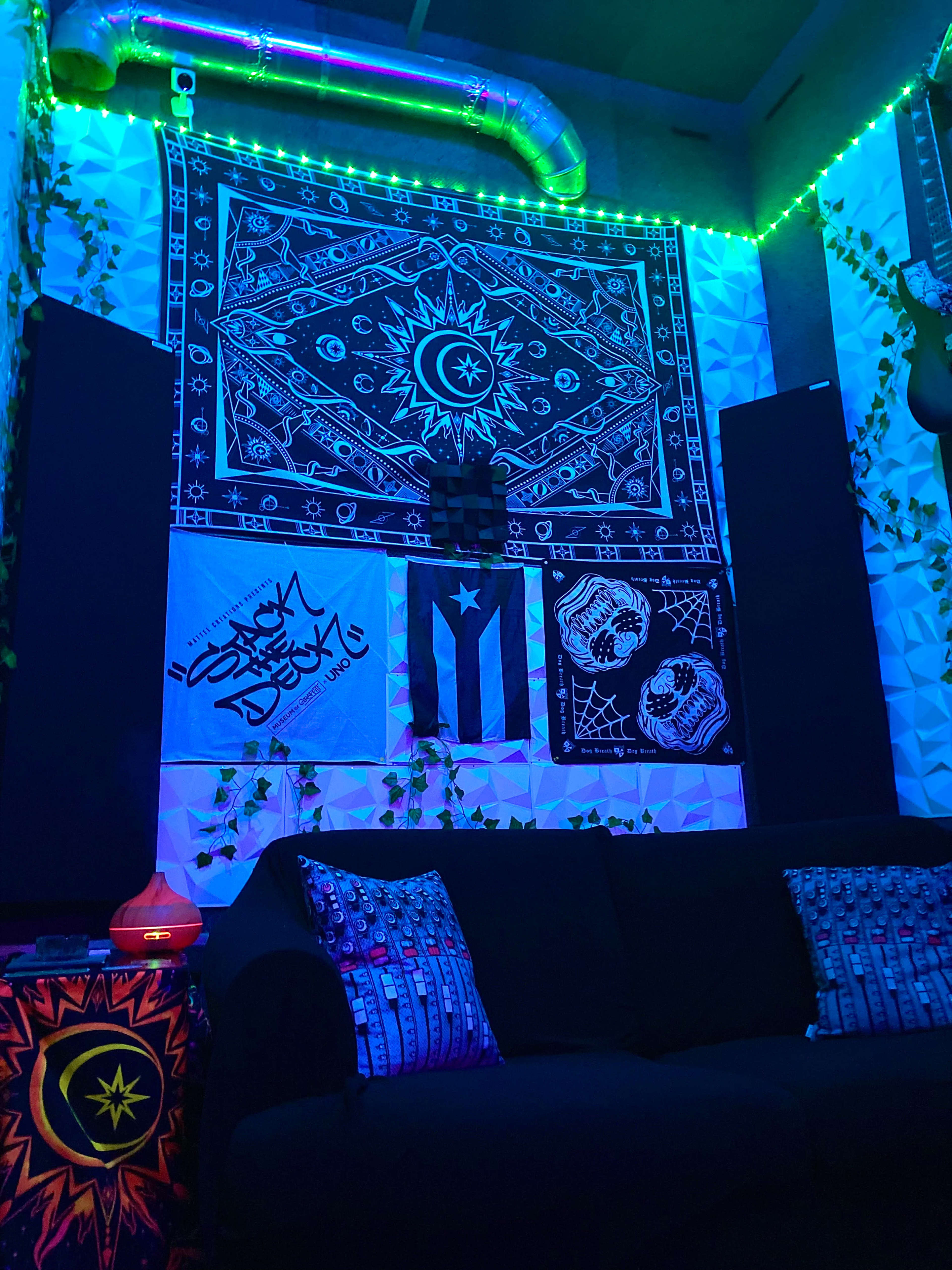 The image shows a cozy room with a dark couch, decorated walls featuring tapestries, and ambient lighting, with plants and musical instruments.
