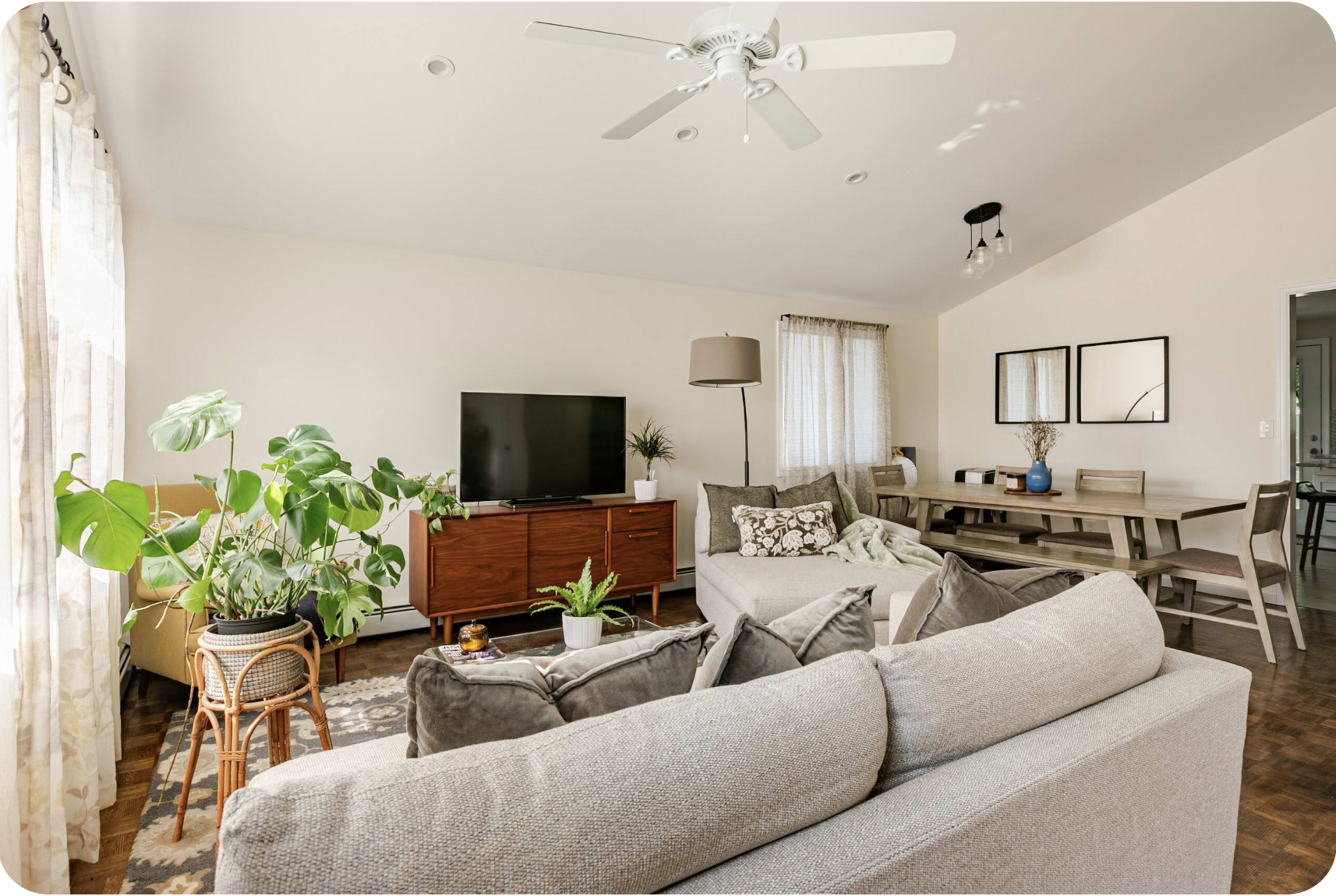 The image shows a modern living room with a large sectional sofa, a coffee table, a TV stand, and a dining area featuring a wooden table and chairs, surrounded by plants and natural light.