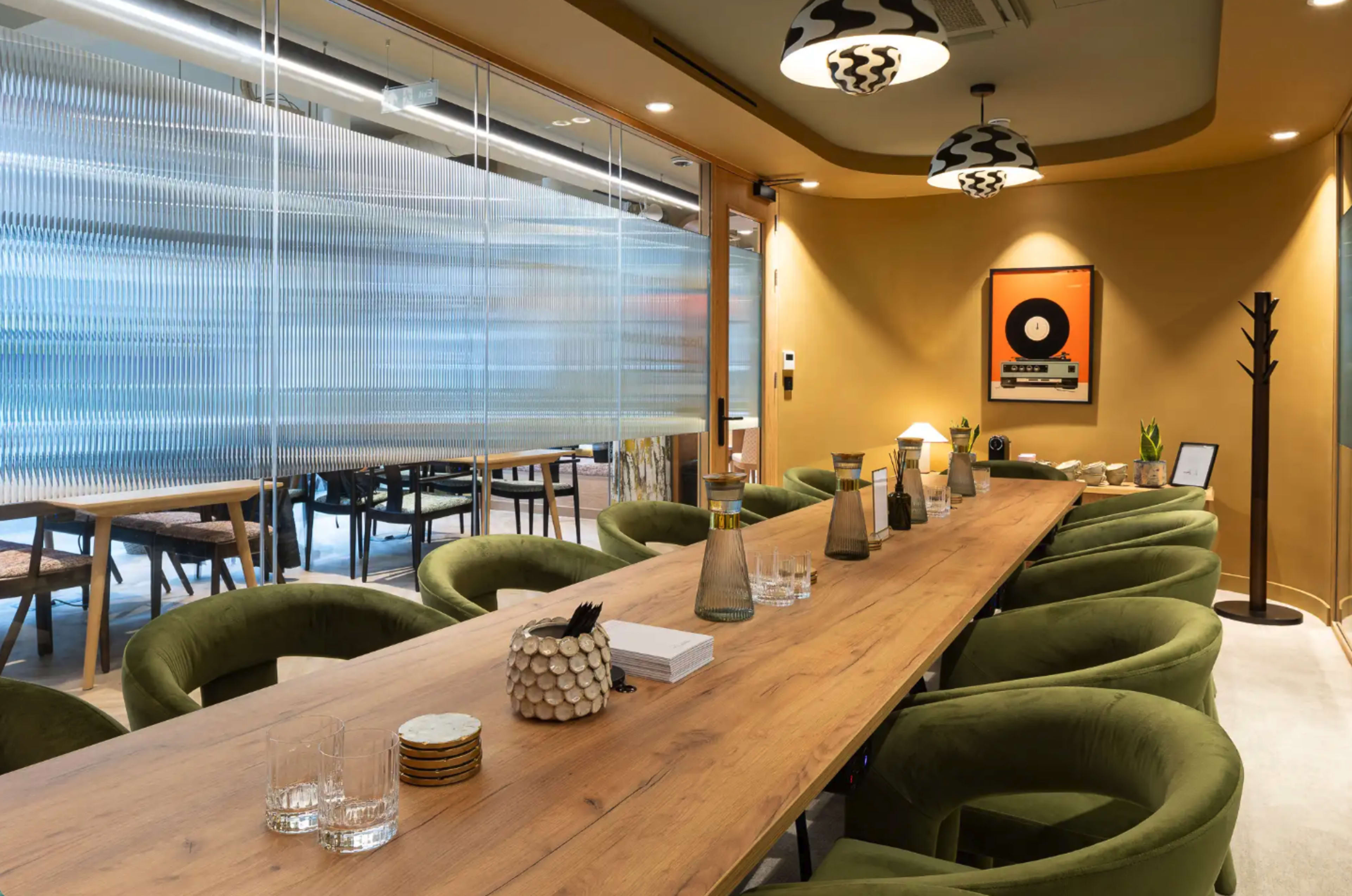 A long wooden table with green upholstered chairs is set in a modern dining area featuring glass partitions and decorative lighting.