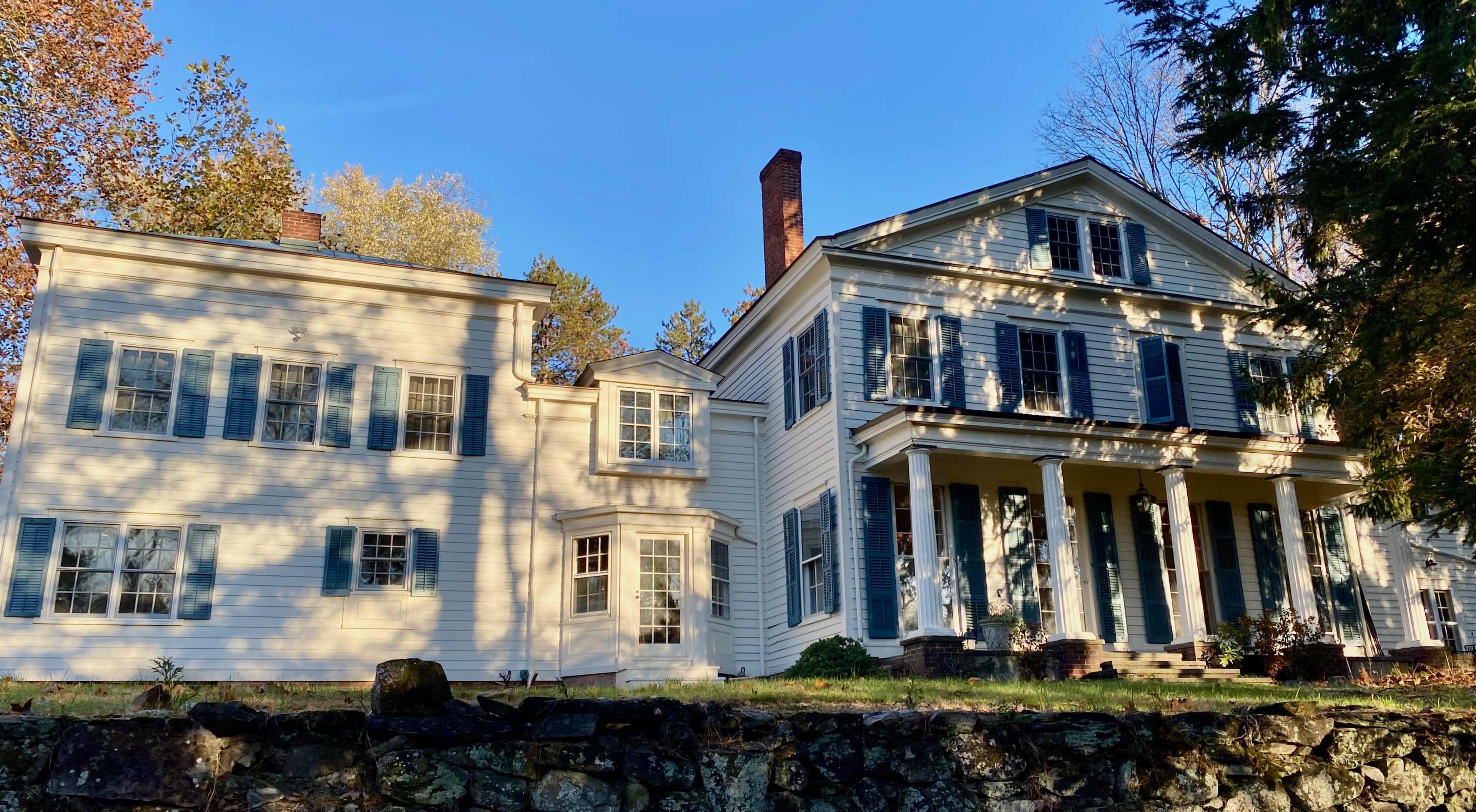 A large two-story white house with blue shutters features a front porch supported by columns and is surrounded by trees and a stone wall.