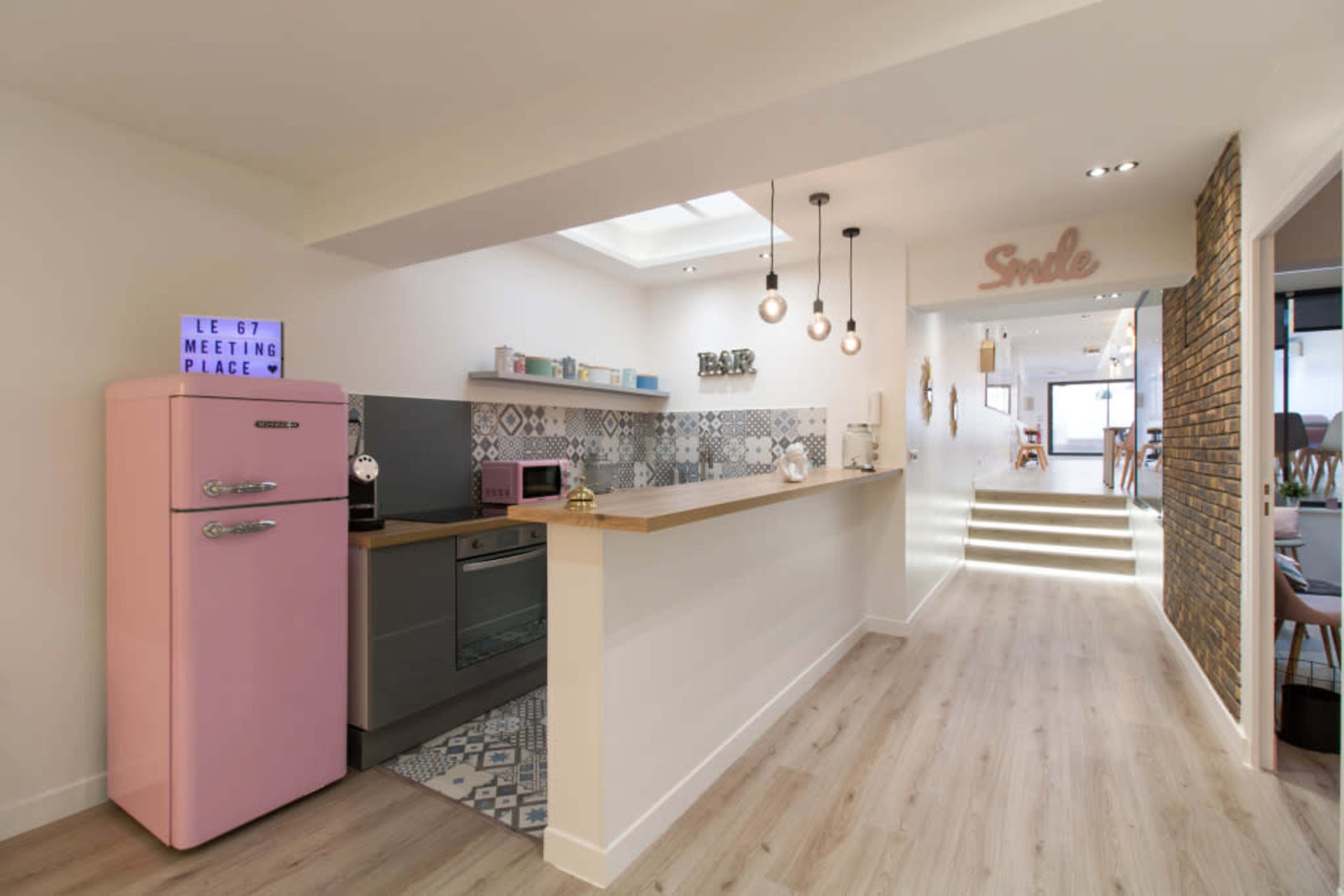 A modern kitchen with a pink refrigerator, a countertop, and decorative tiles, leading to a well-lit hallway with stairs.