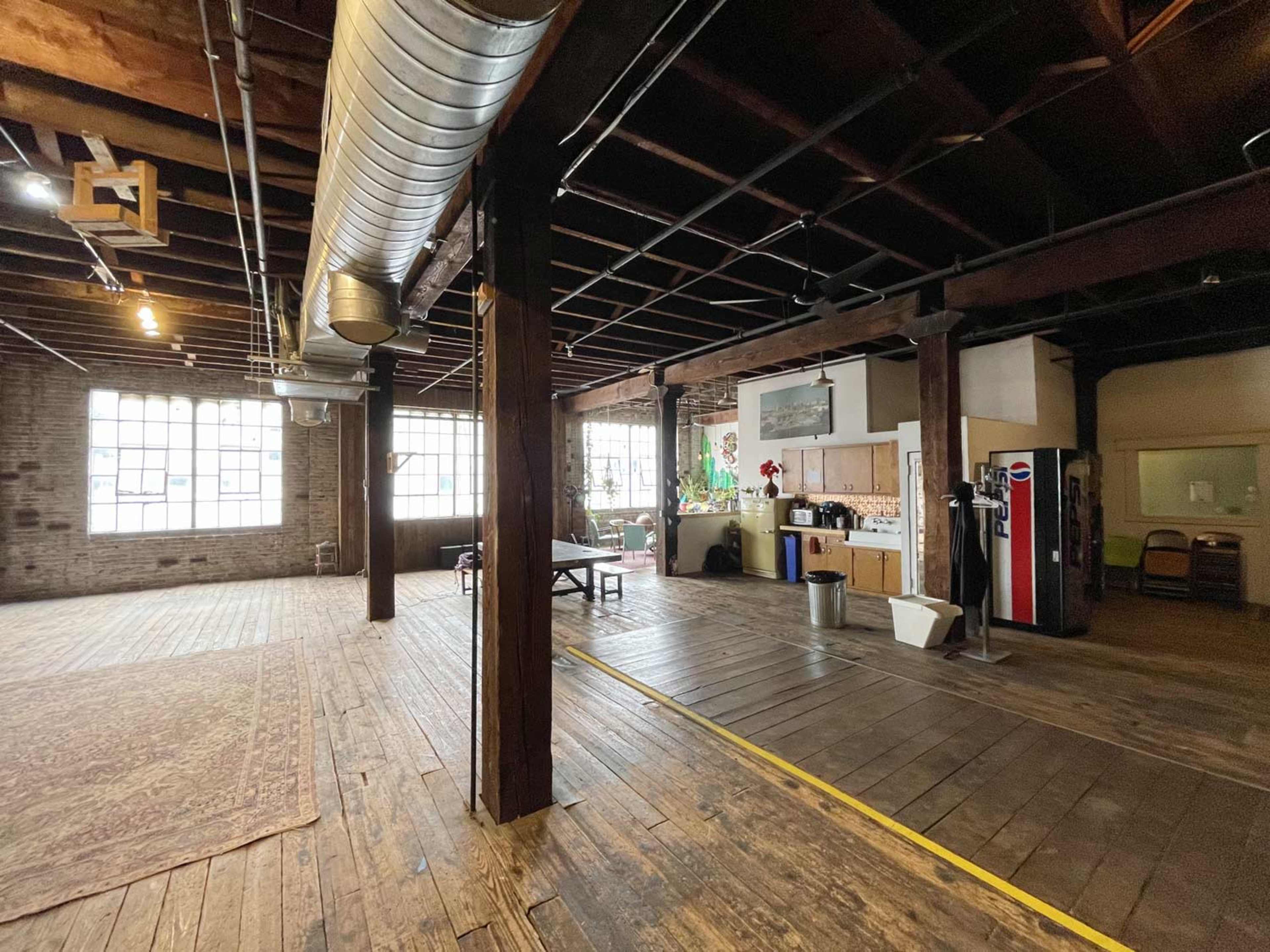 The image shows a spacious, industrial-style room with wooden flooring, exposed beams, and large windows, featuring a kitchen area and a table in the center.