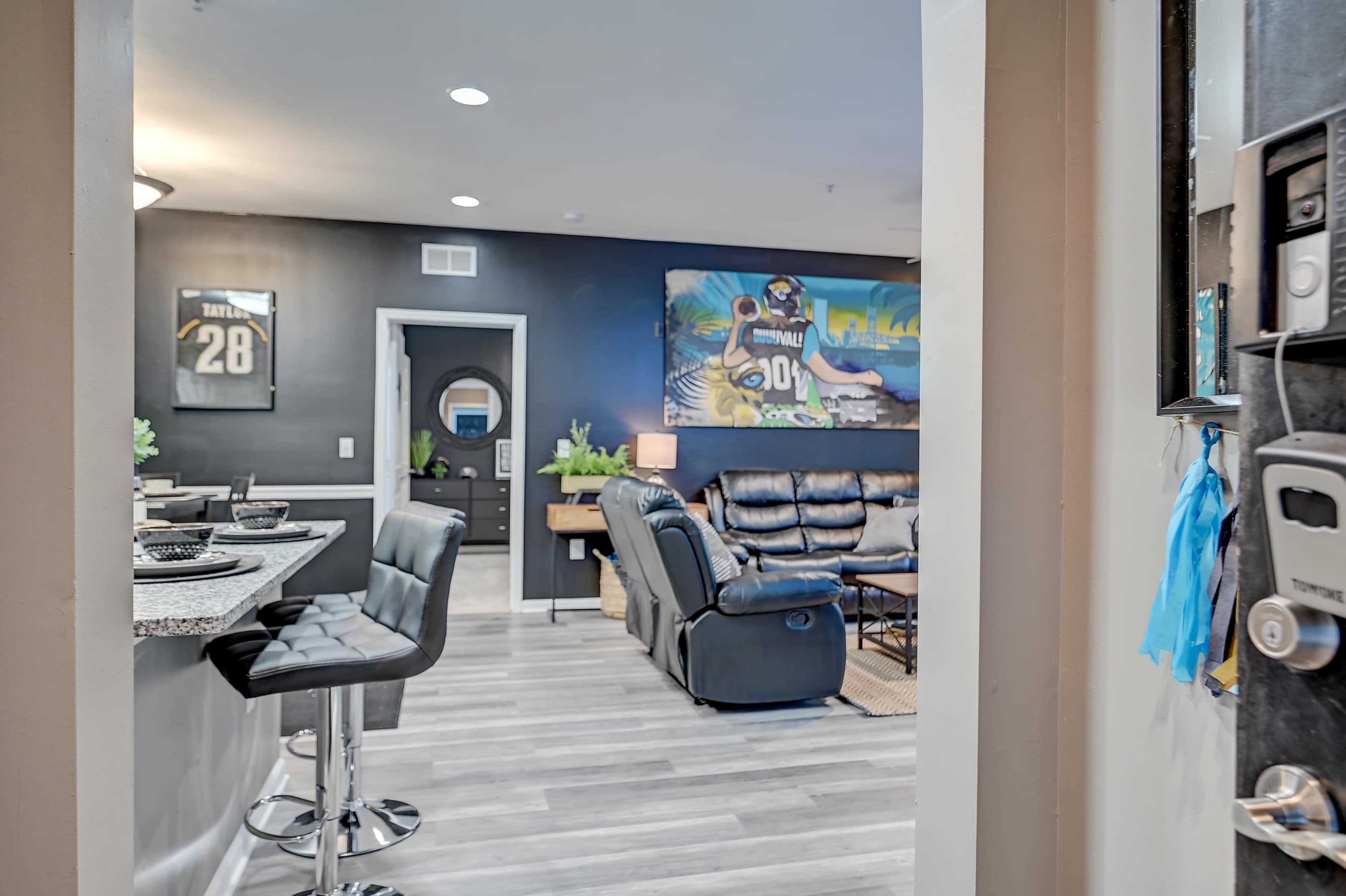 The image shows an open living area with a kitchen bar and seating area, featuring black leather furniture and a large wall painting.