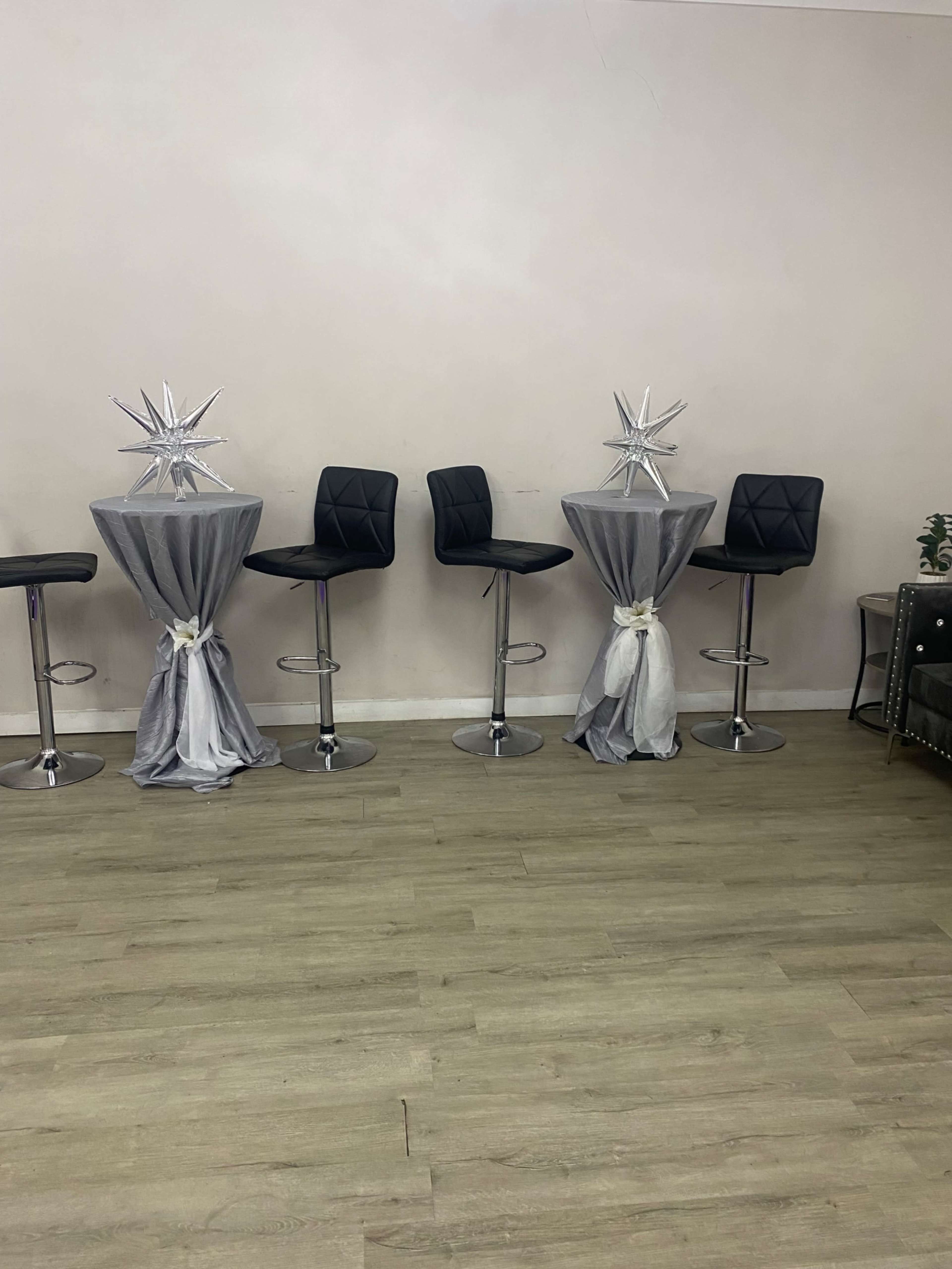 Several high-top tables with gray tablecloths and black bar stools arranged in a room with light wood flooring.
