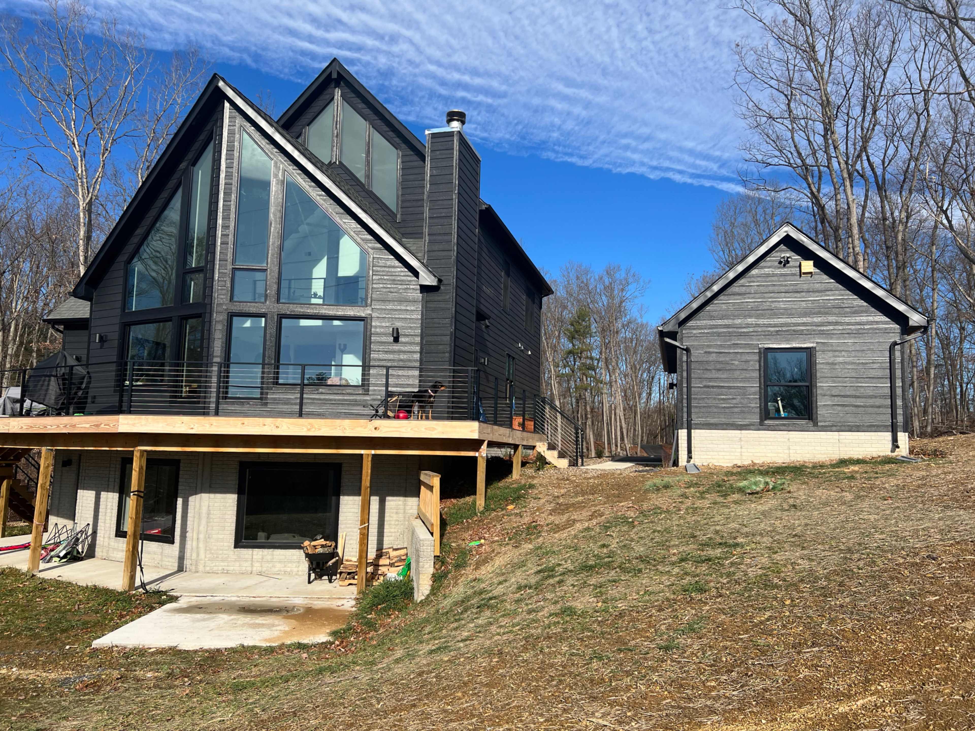 The image shows a modern, two-building residential property with large windows and a contrasting smaller structure situated in a wooded area.