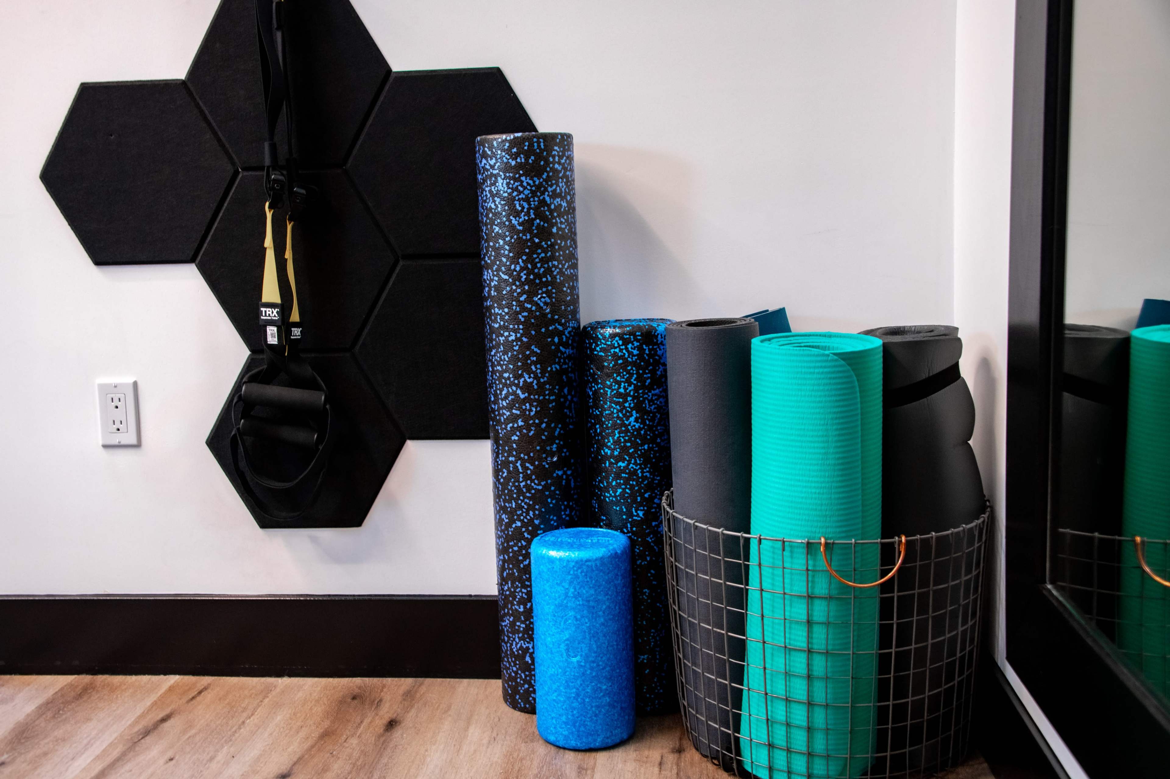 A wall-mounted exercise strap alongside a collection of various yoga mats and foam rollers stored in a wire basket near a mirror.