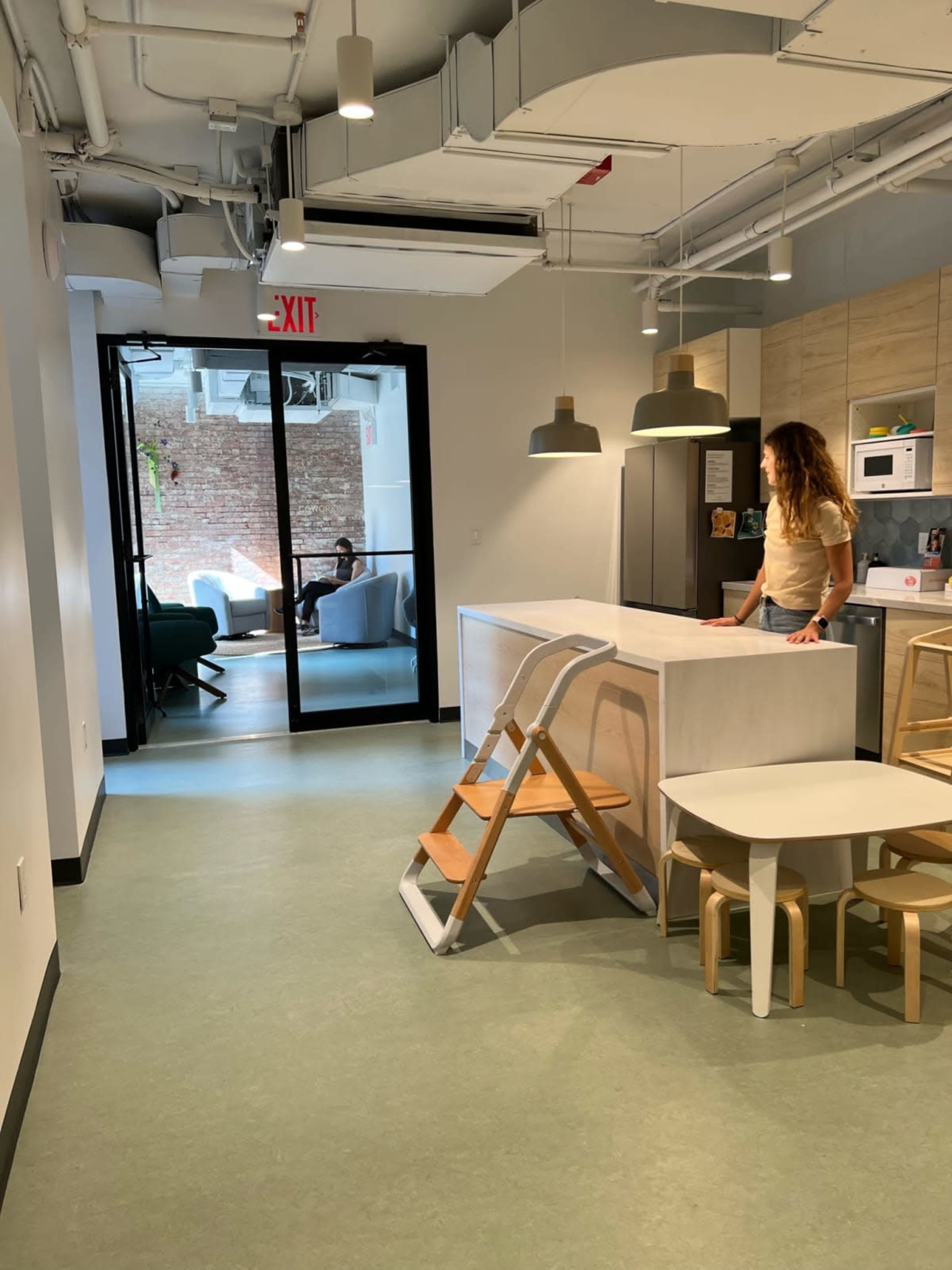 A kitchen area with a person standing by a counter, overlooking a doorway that leads to a seating area with a brick wall in the background.