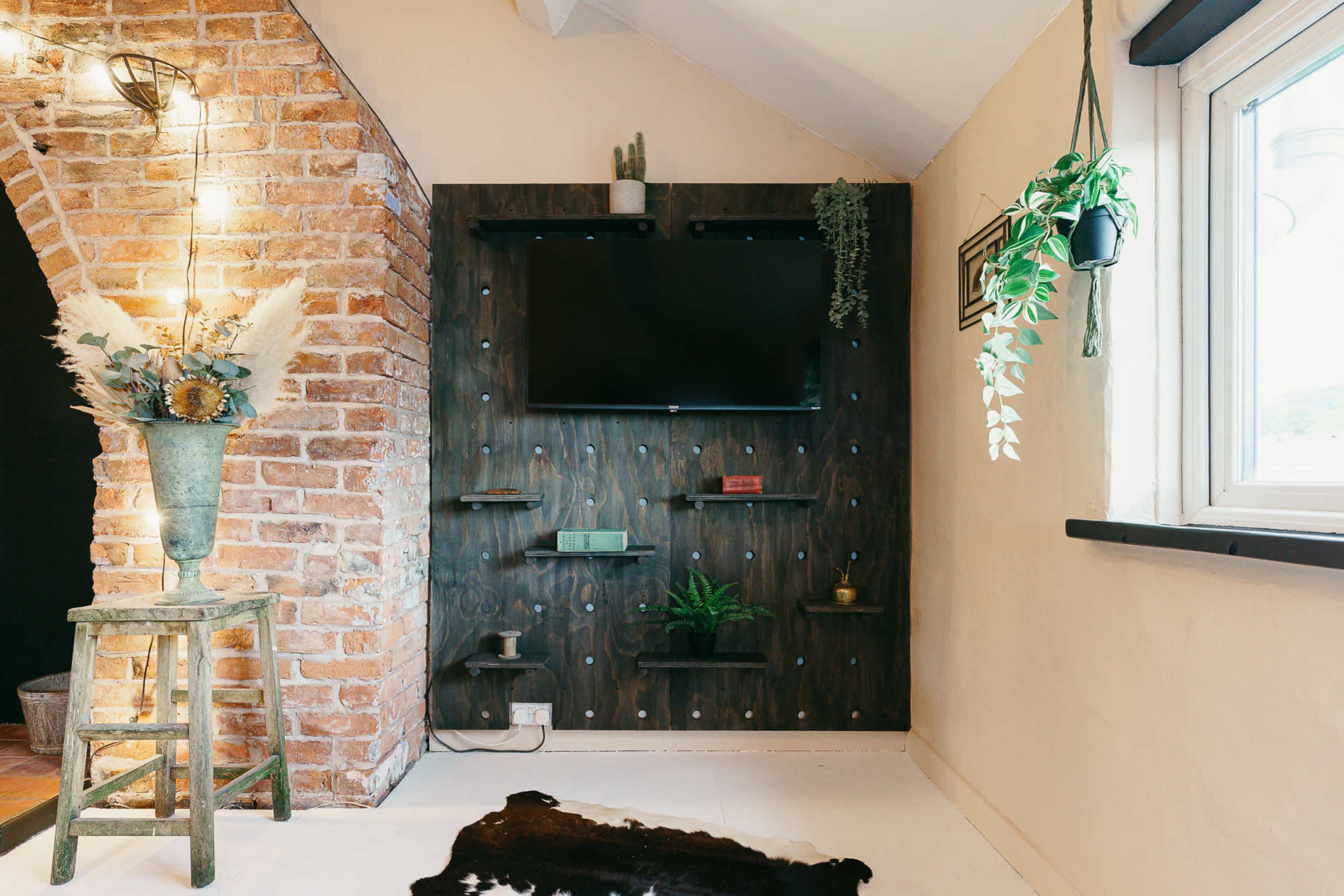 The image shows a cozy corner of a room with a wall-mounted TV, a dark wooden shelving unit, and a small wooden stool beside decorative plants and a cowhide rug.