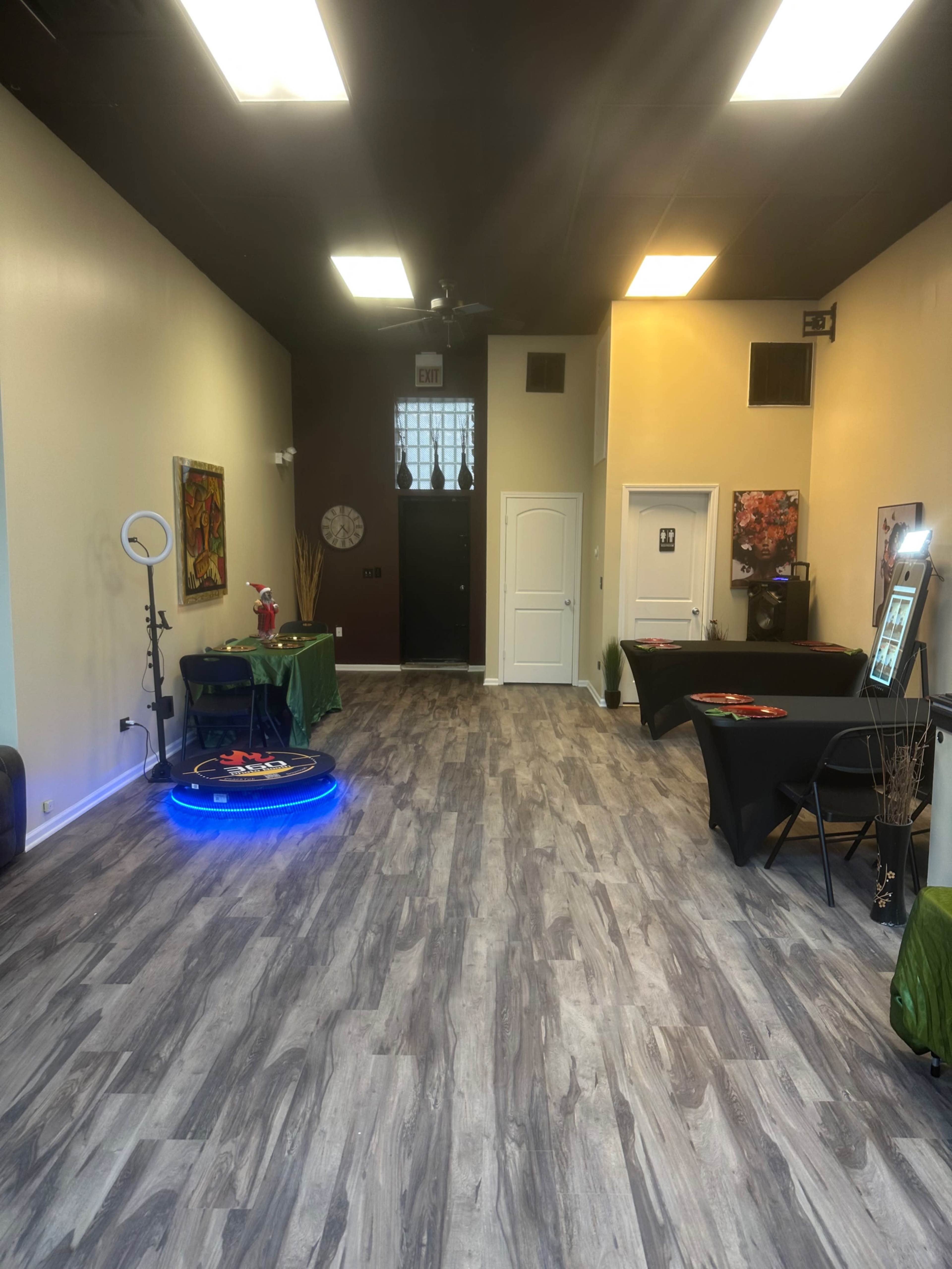 The image shows a spacious indoor area with wooden-like flooring, featuring various tables and decorations, including a ring light and a hoverboard in the center.
