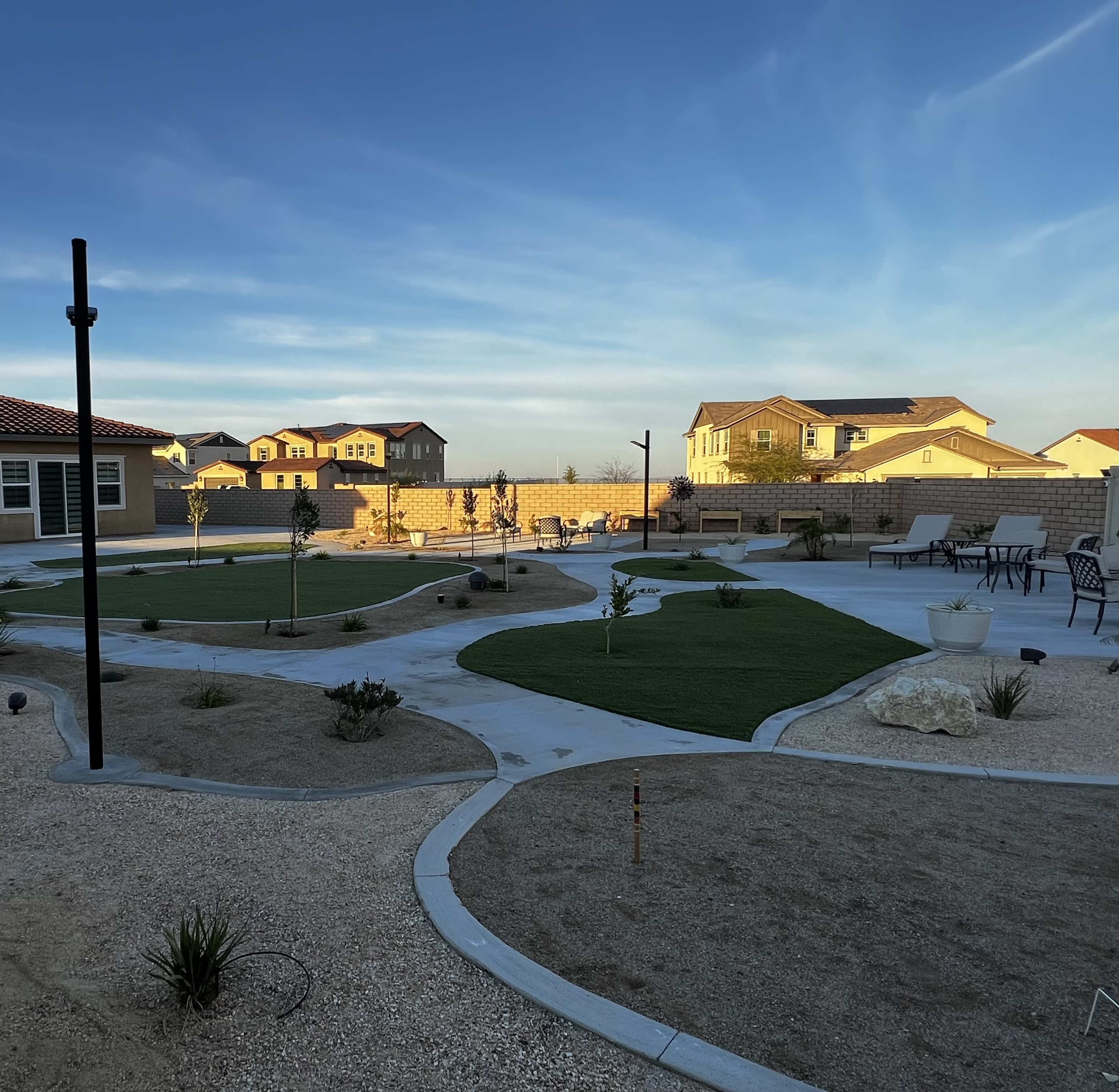 The image shows a landscaped outdoor area with patchy grass, stone paths, and seating arrangements, surrounded by residential buildings.