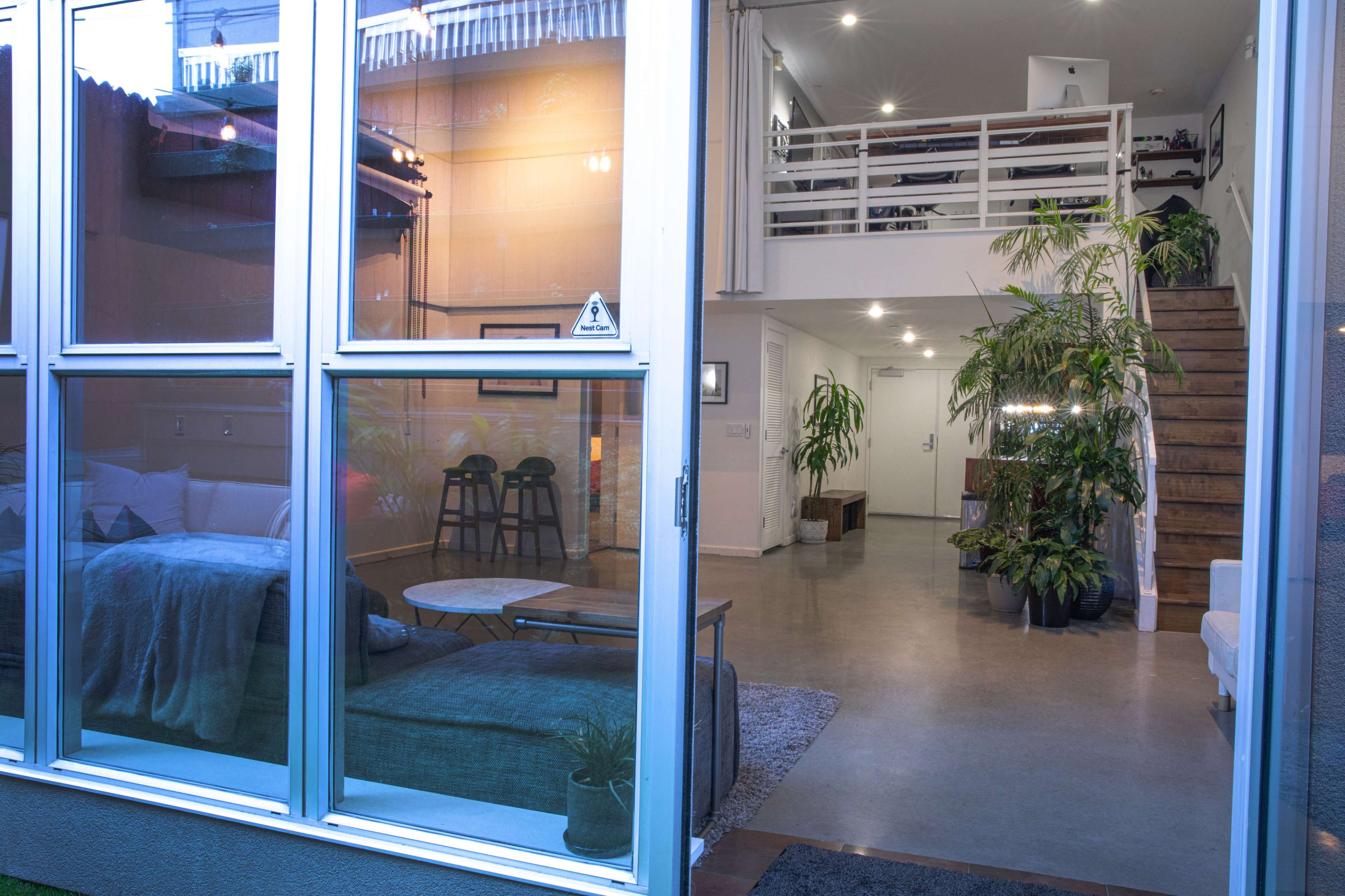 A modern, open-concept interior with large glass windows, a staircase leading to an upper level, and various indoor plants.