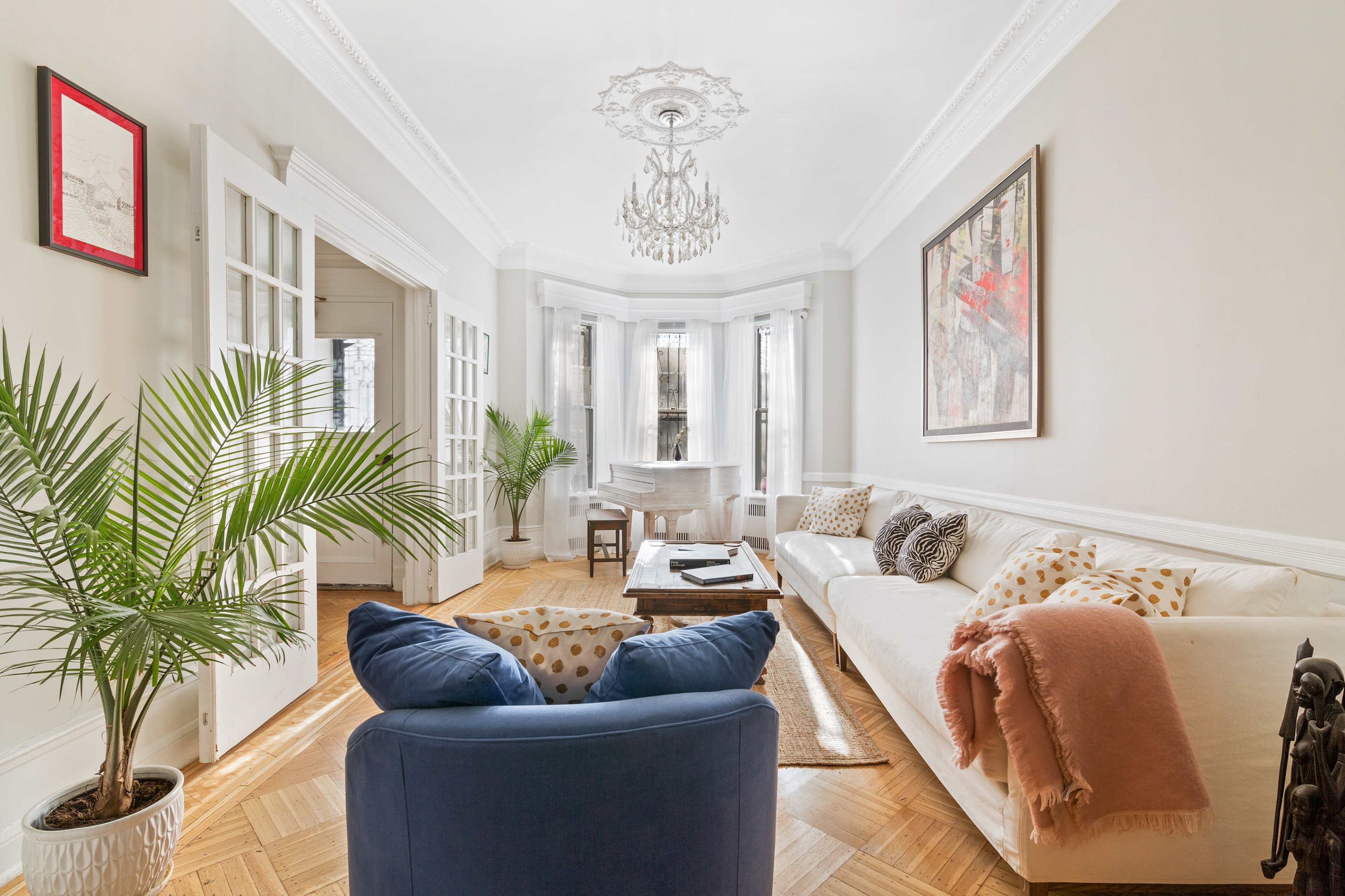 A bright living room features a white sofa with patterned cushions, a blue armchair, a plant in a pot, and a grand piano in the corner.