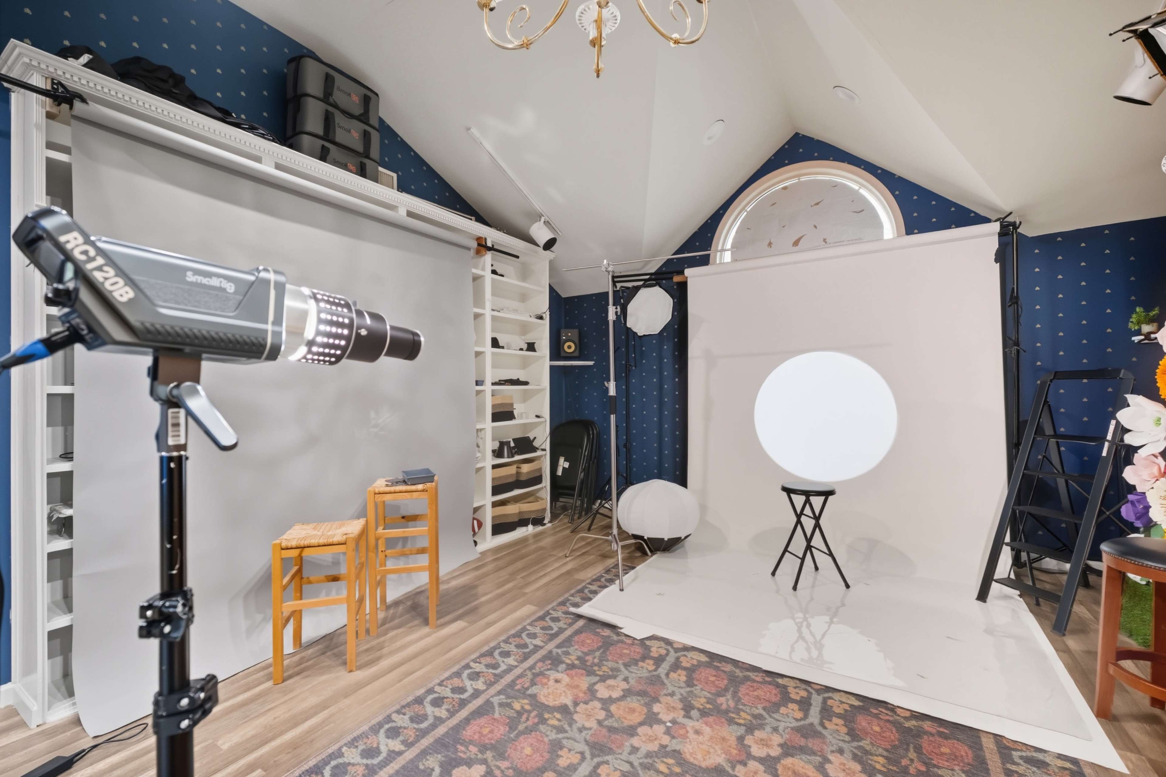 The image shows a photography studio featuring a backdrop setup, a light stand with a flash, and a small chair on a white floor surrounded by decorative elements.