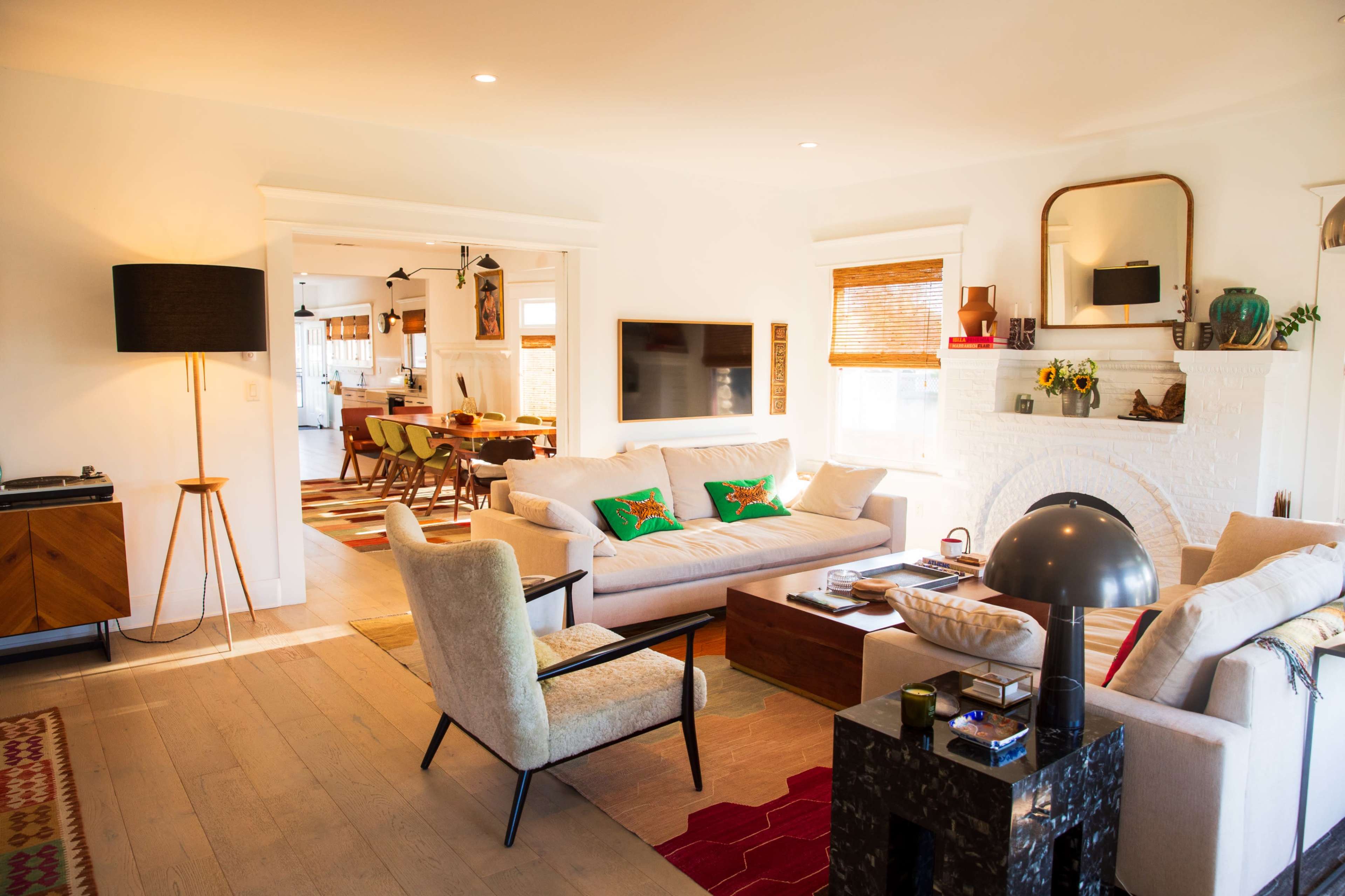 The image shows a bright, modern living room with a light-colored sofa, a small coffee table, and a fireplace, featuring an open layout that connects to a dining area in the background.