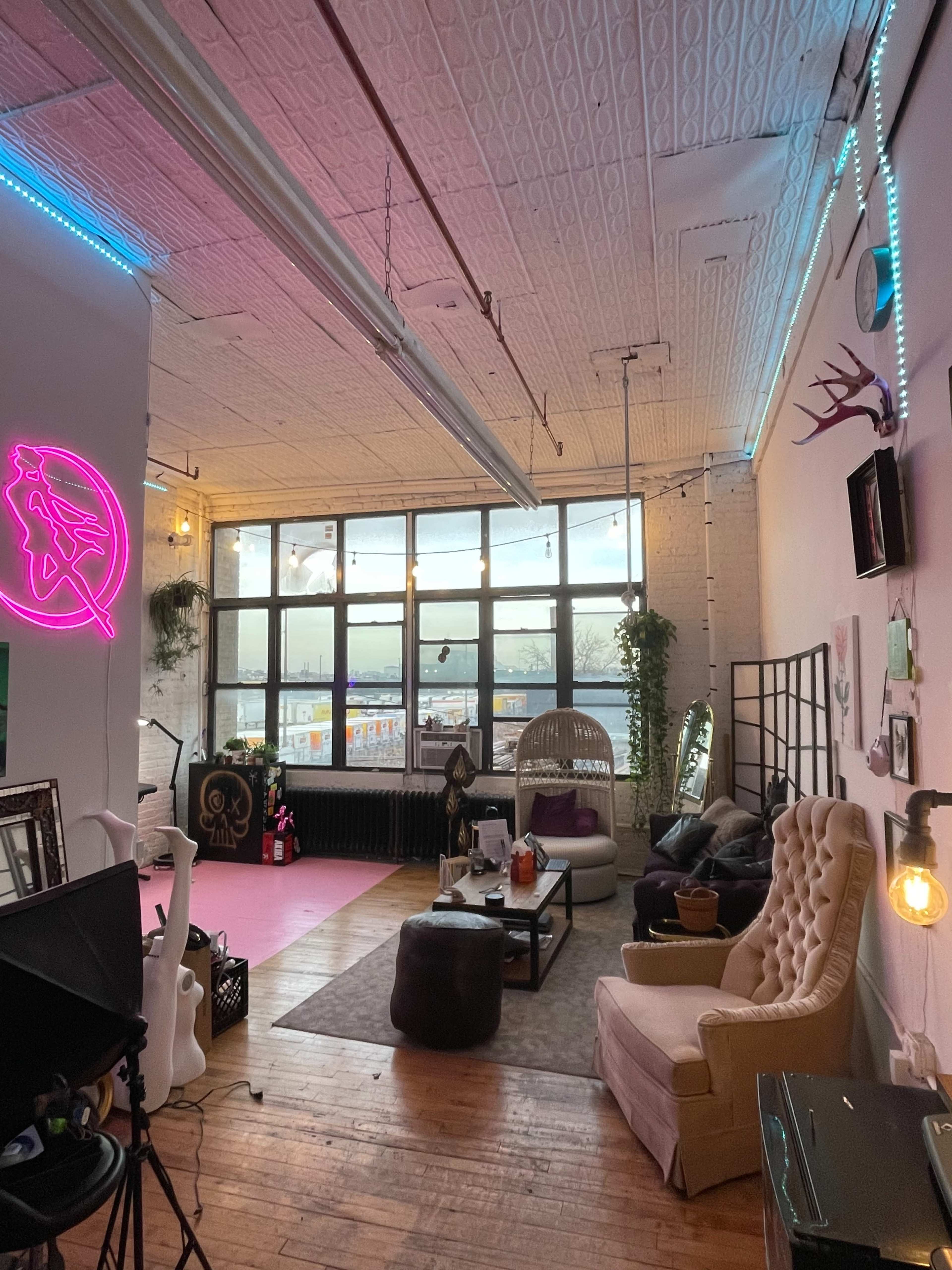 The image shows a spacious, well-lit room with large windows, featuring a mix of modern and cozy furniture, decorative plants, and neon lights.