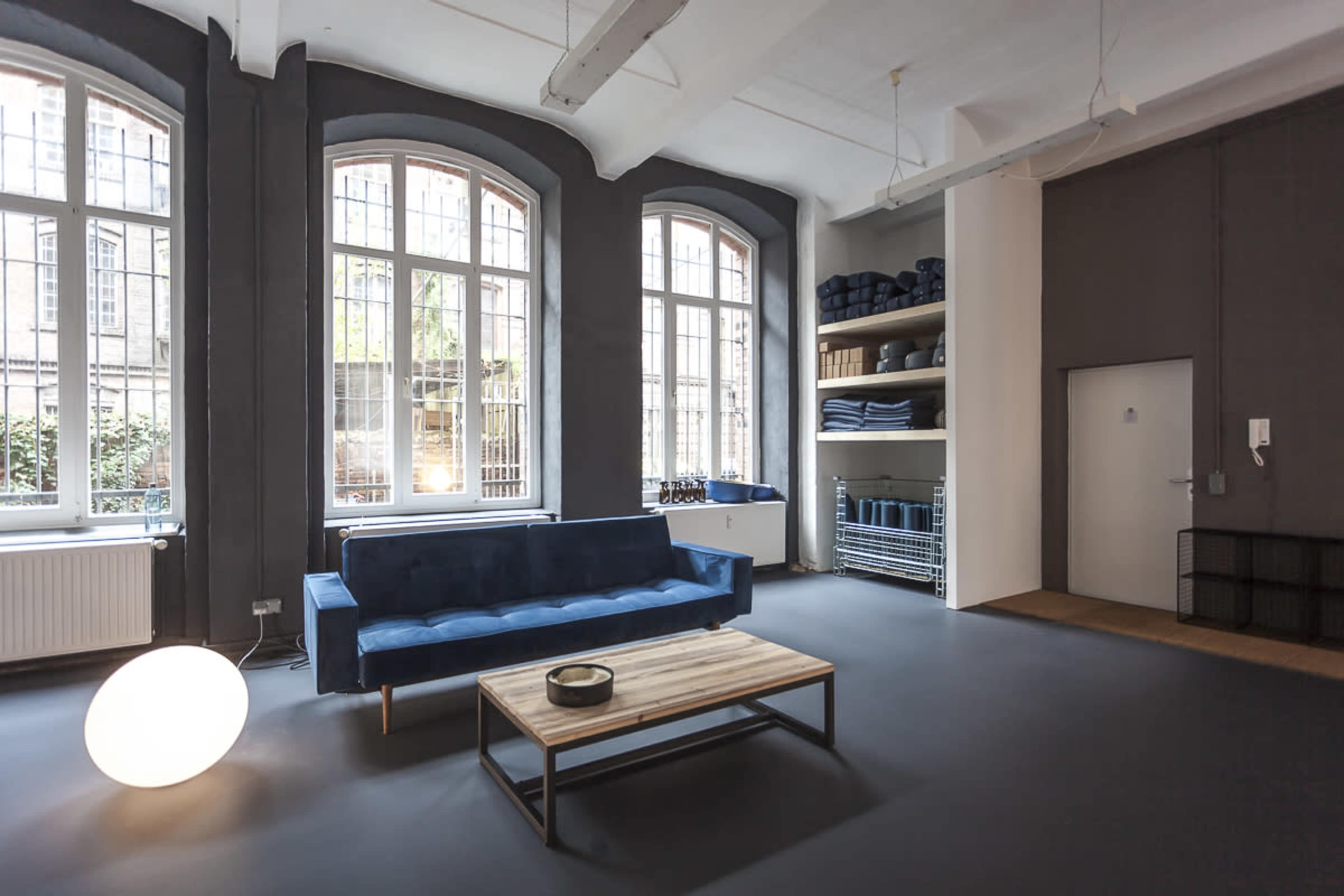 The image shows a spacious room with large windows, a blue sofa, a wooden coffee table, and shelves piled with neatly folded towels.