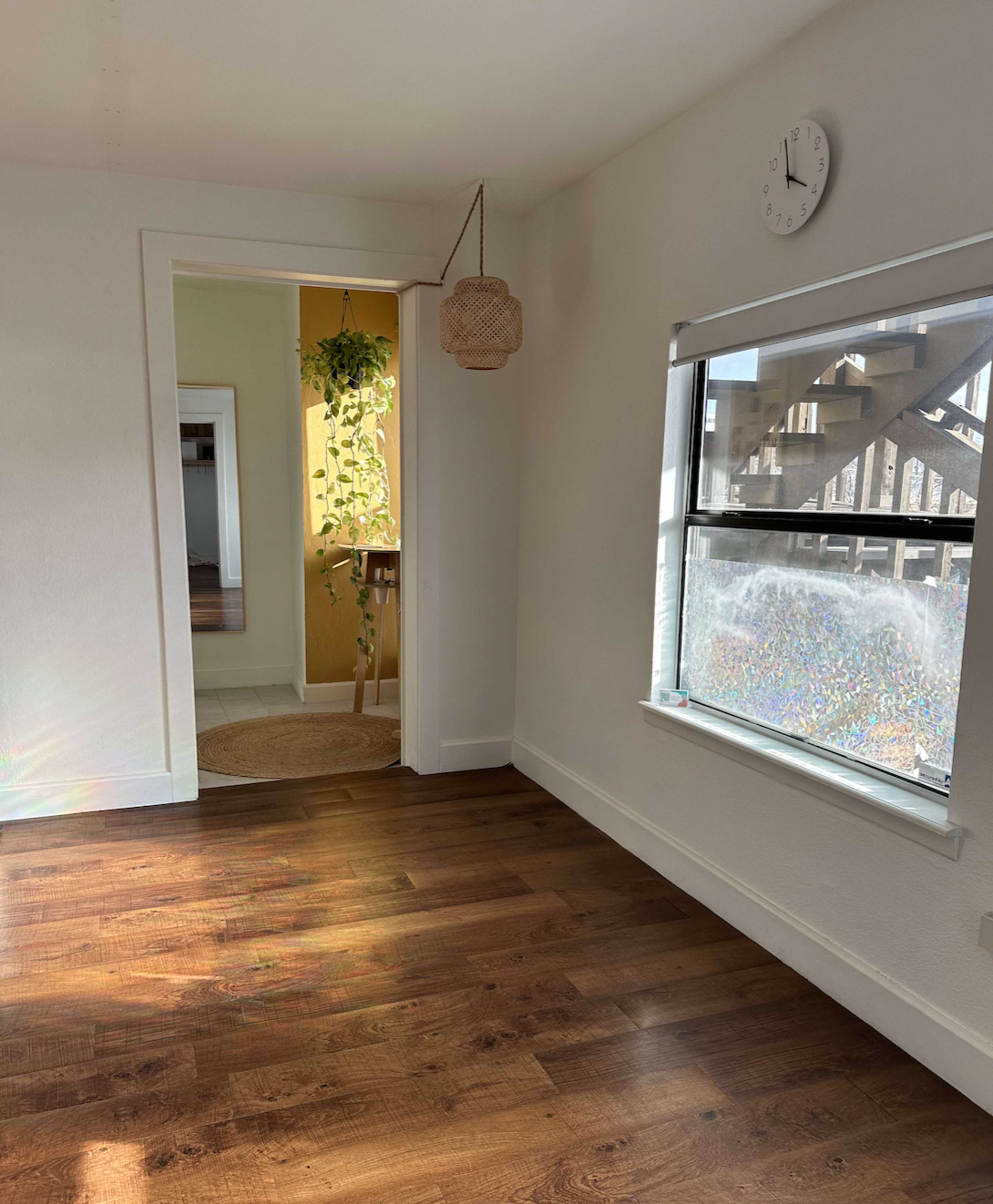 The image shows a bright room with light-colored walls, a wooden floor, a window, and an open door leading to a yellow-walled area with a plant hanging.