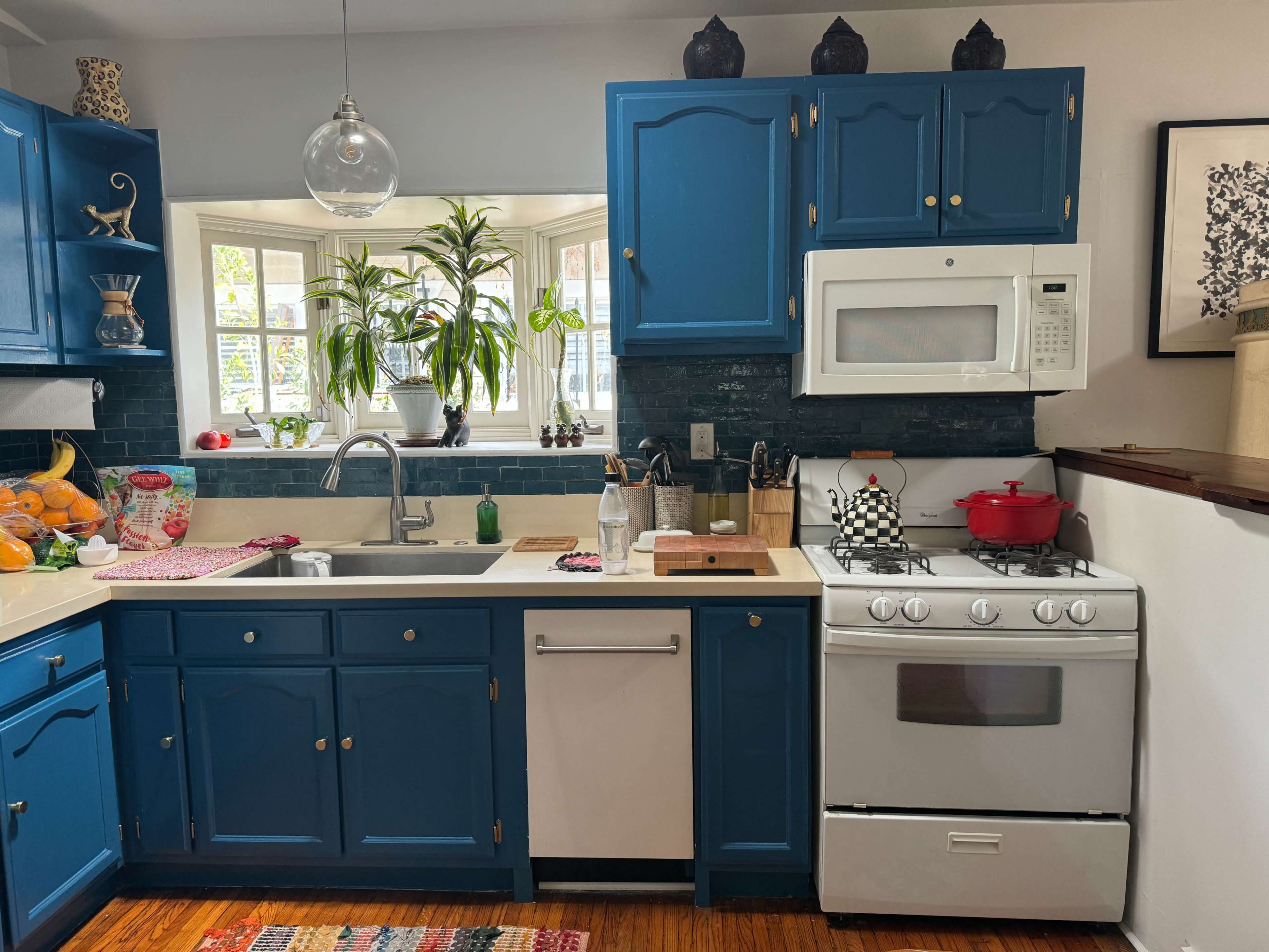 A kitchen features blue cabinetry, a white stove, and a sink beneath a window with potted plants.