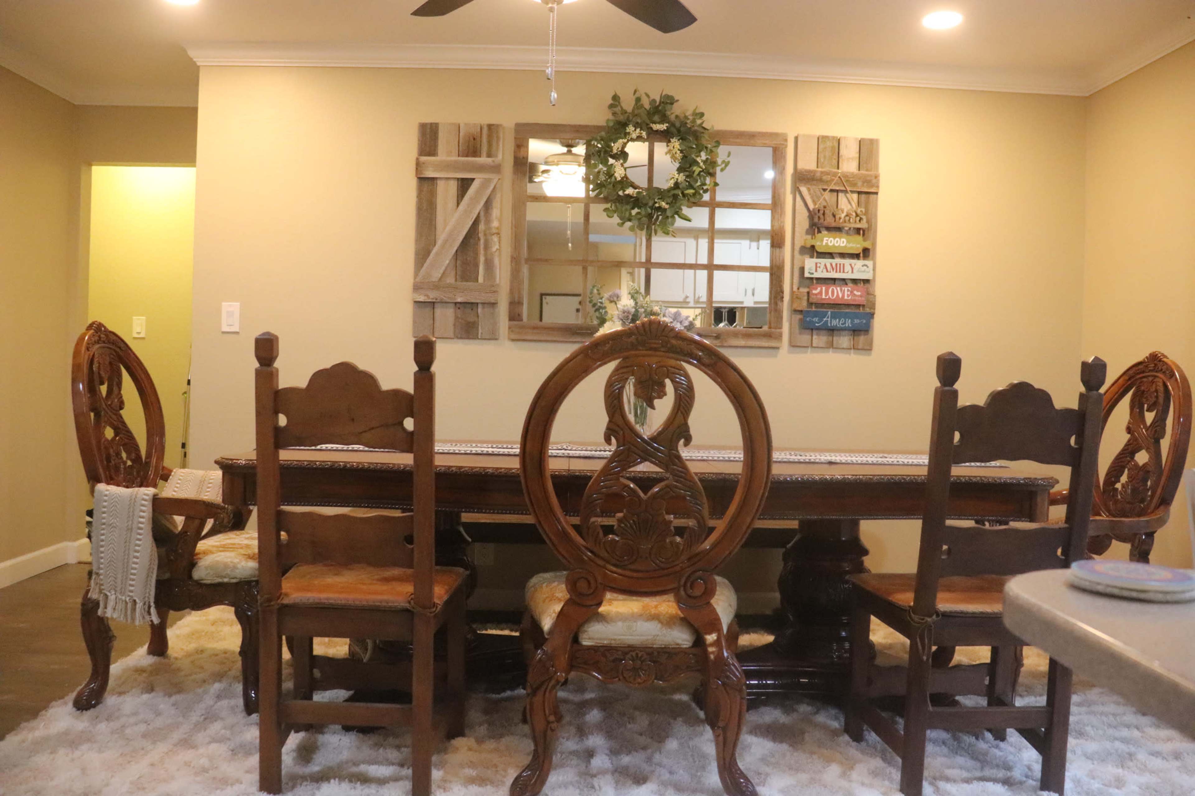 A dining room features a large wooden table surrounded by six ornate chairs, with a decorative mirror and wall art displayed in the background.