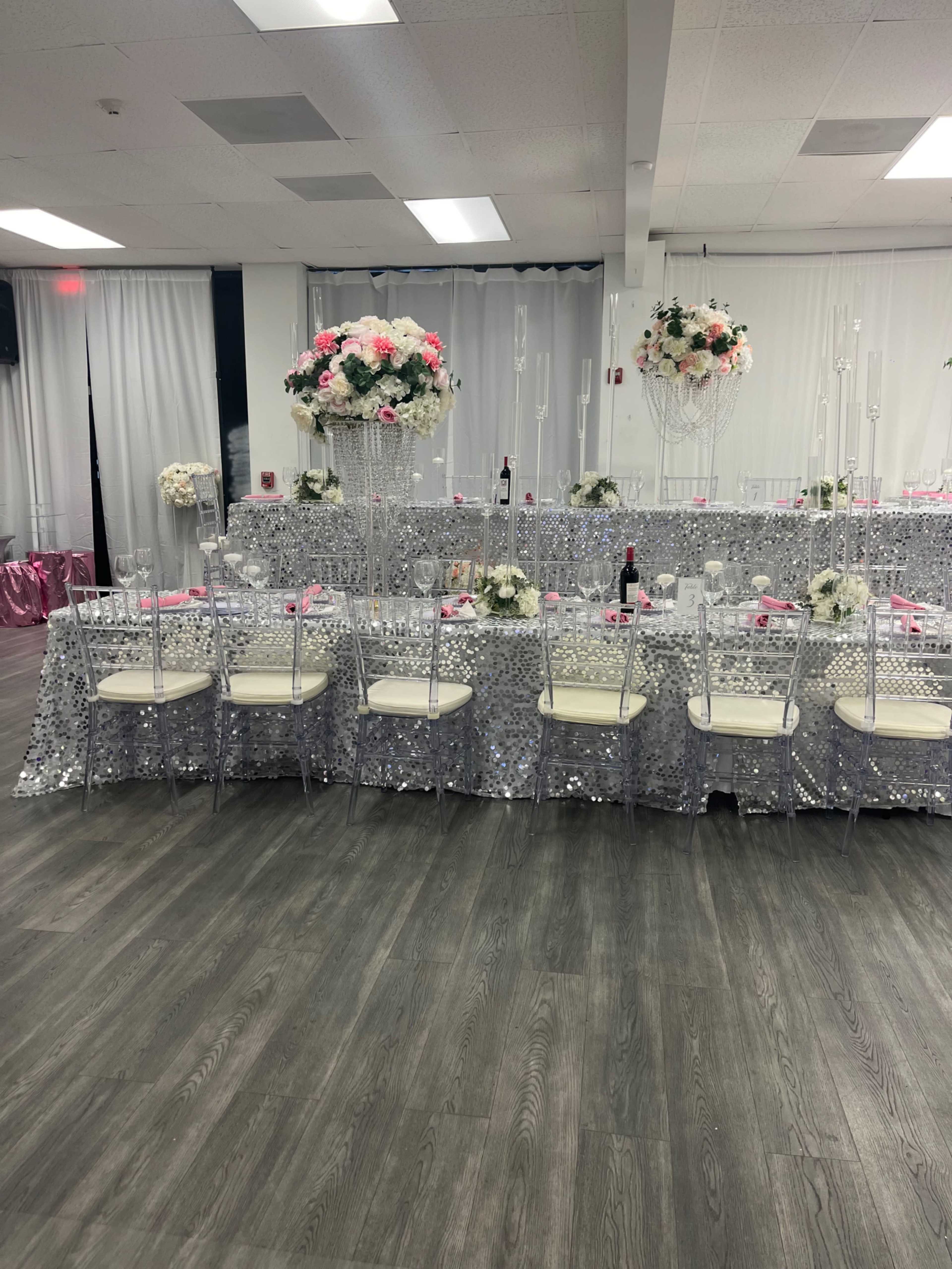 A banquet hall setup with several tables covered in silver sequin tablecloths, adorned with floral centerpieces and white chairs arranged around them.