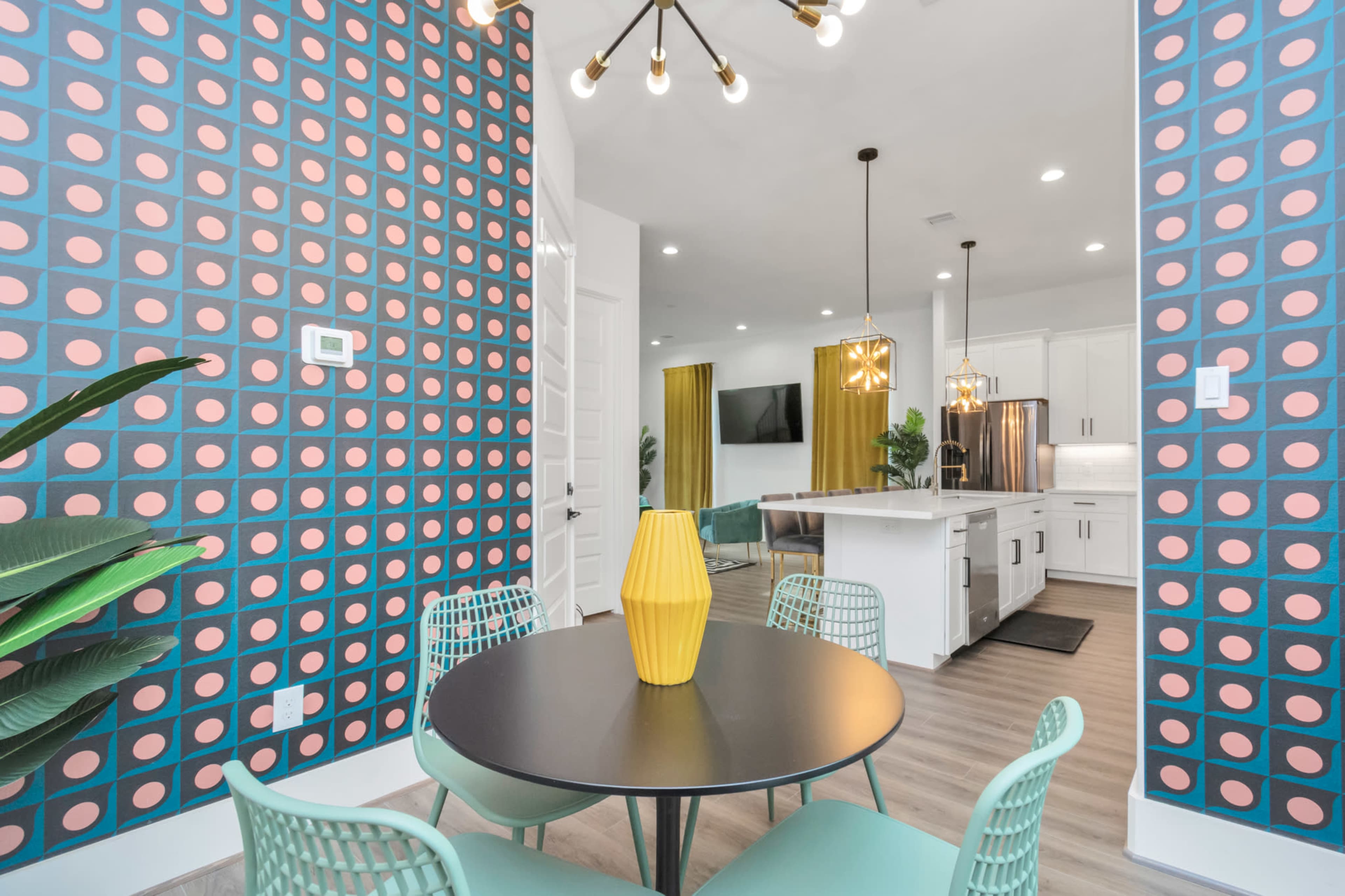 A dining area features a round black table surrounded by mint green chairs, against a backdrop of patterned wallpaper and a modern kitchen visible in the background.