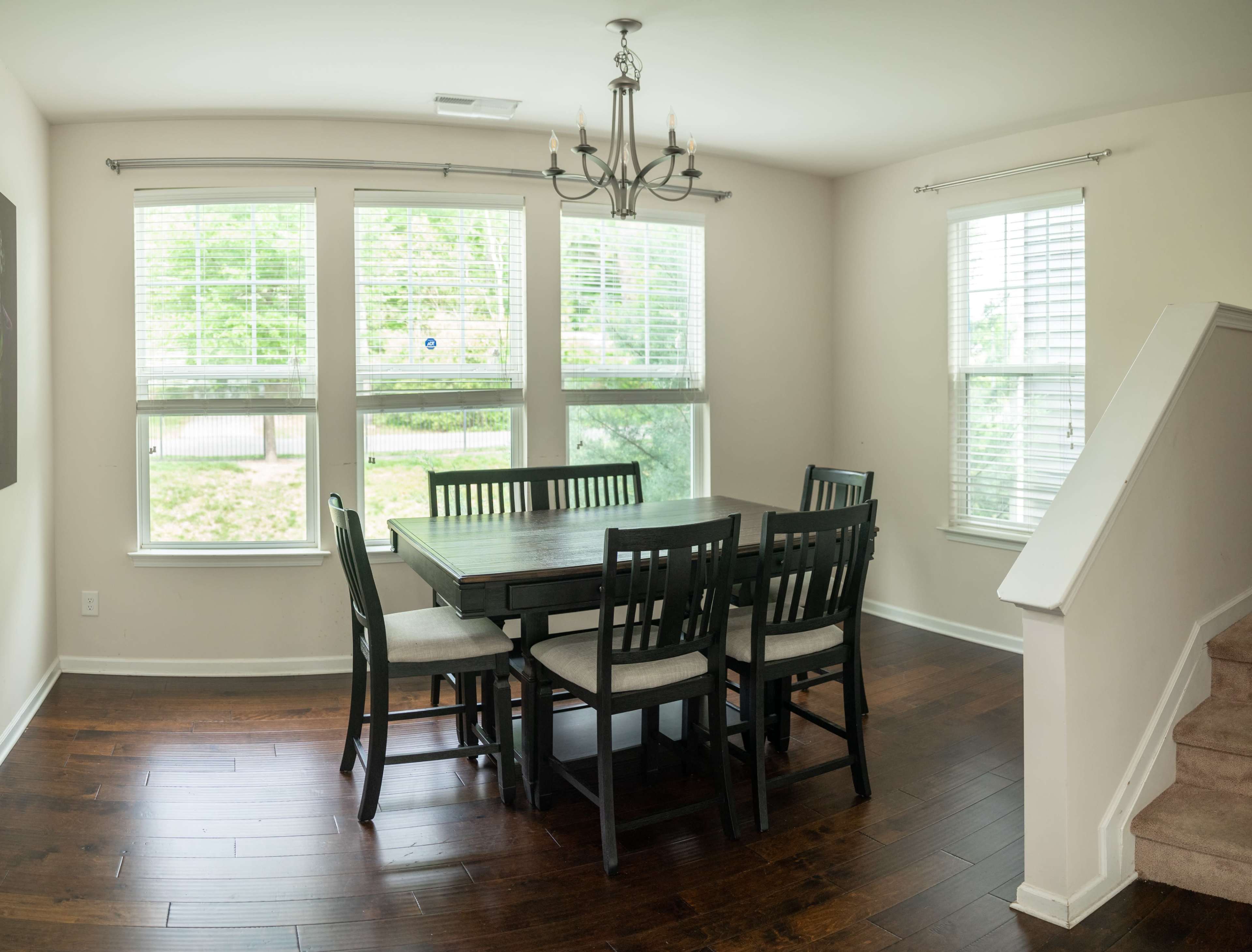 A dining area features a rectangular table surrounded by six chairs, with large windows providing natural light and a staircase at the side.