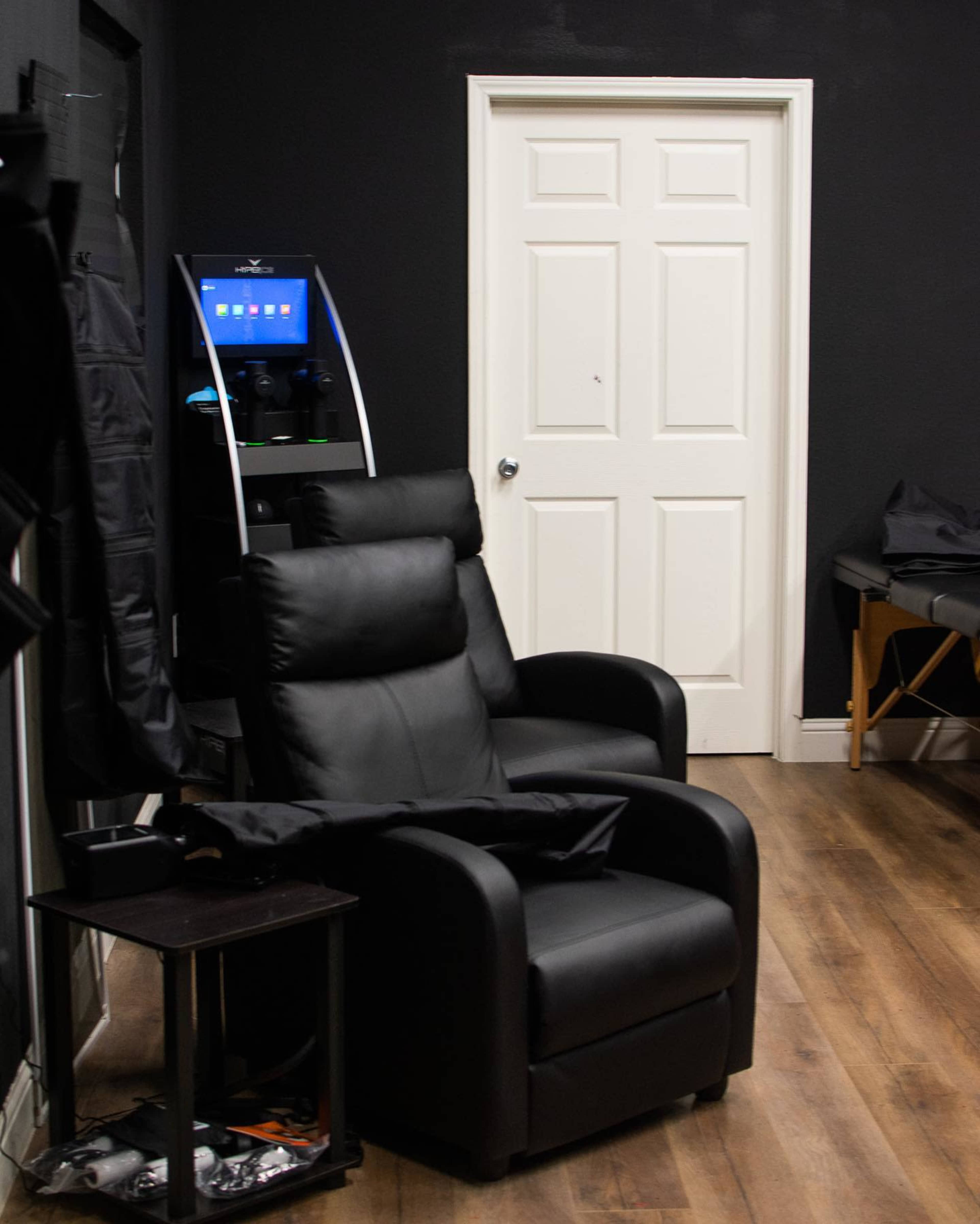 Two black reclining chairs in a room with dark walls, a door, and a beverage station with a touchscreen in the corner.