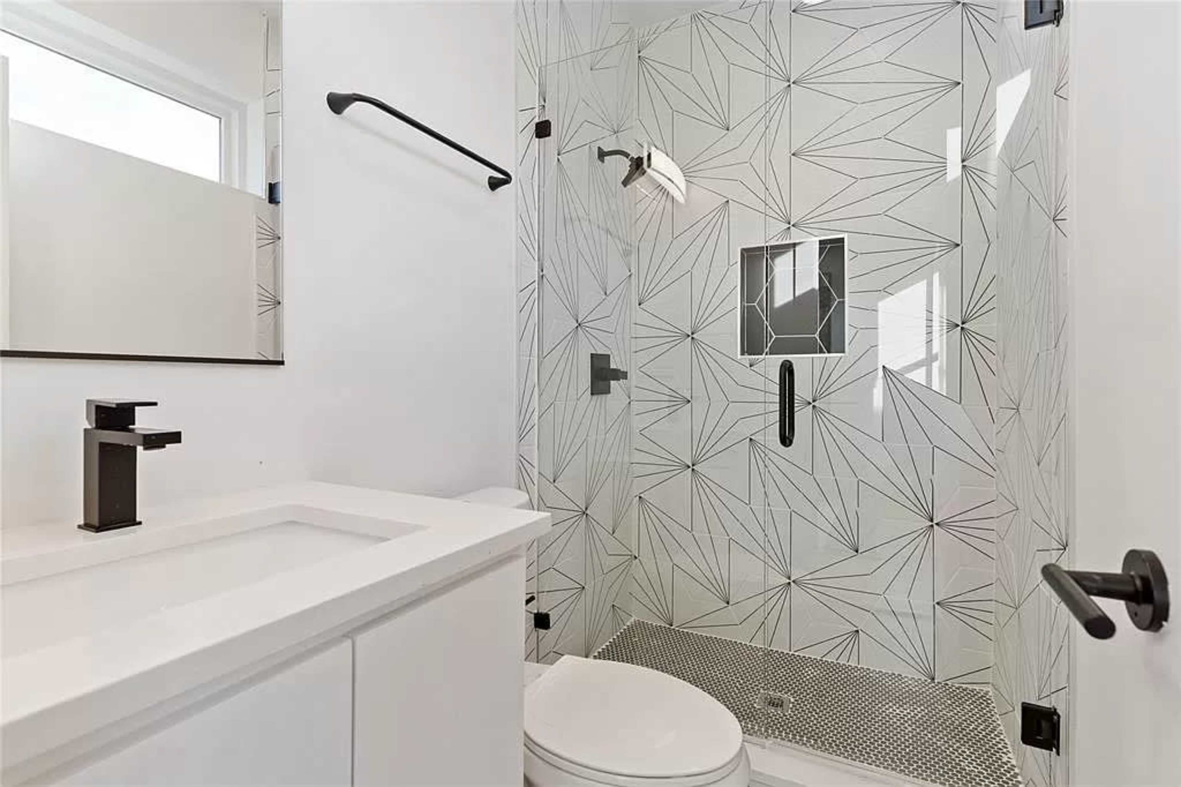 A modern bathroom with a glass shower featuring geometric patterns, a wall-mounted mirror, and a white sink with black fixtures.